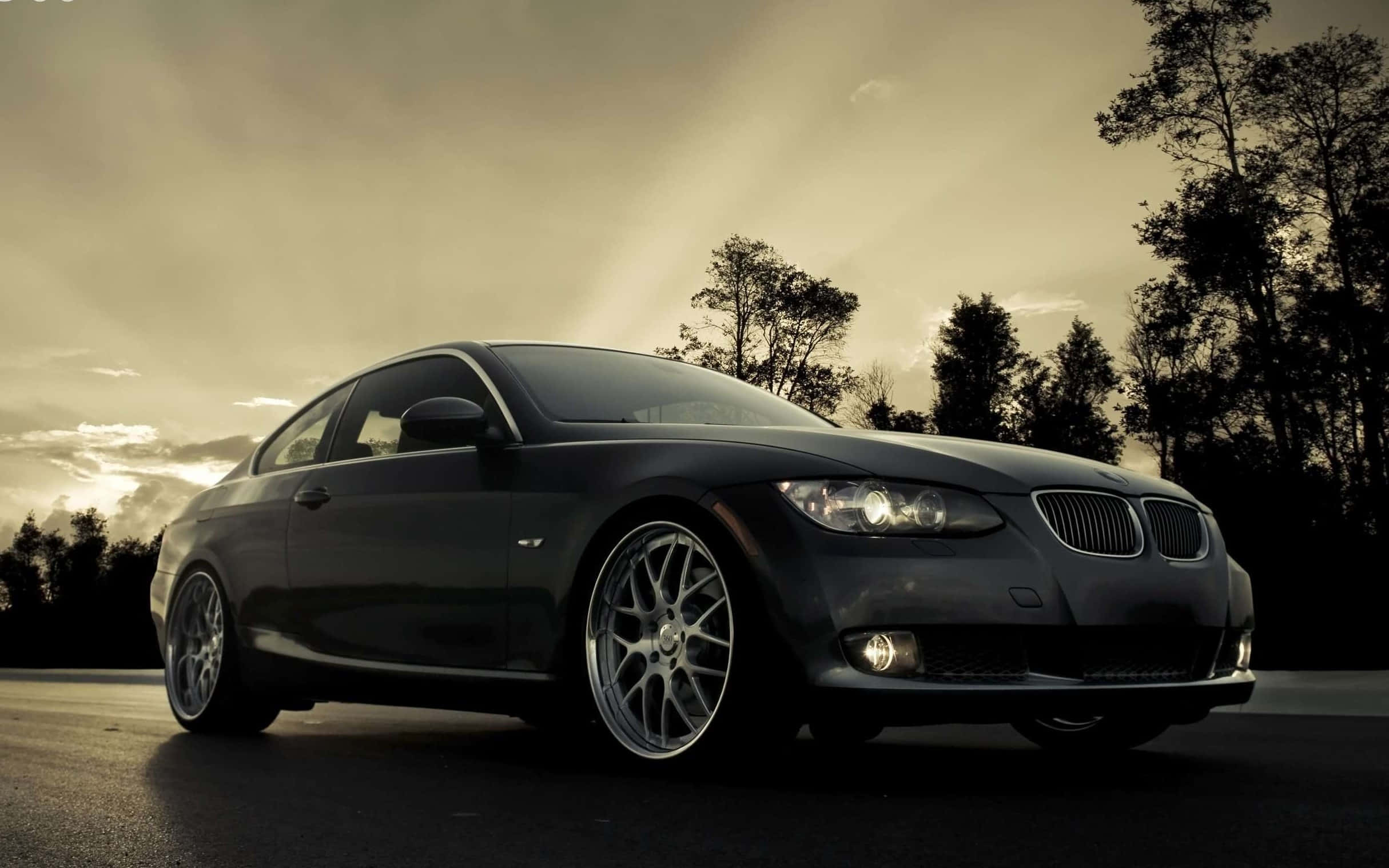 Bmw 328 桌布 壁紙 — 一輛時尚的黑色BMW雙門跑車在金色夕陽下，於一條兩旁種滿深色樹木的寧靜道路上閃閃發光。