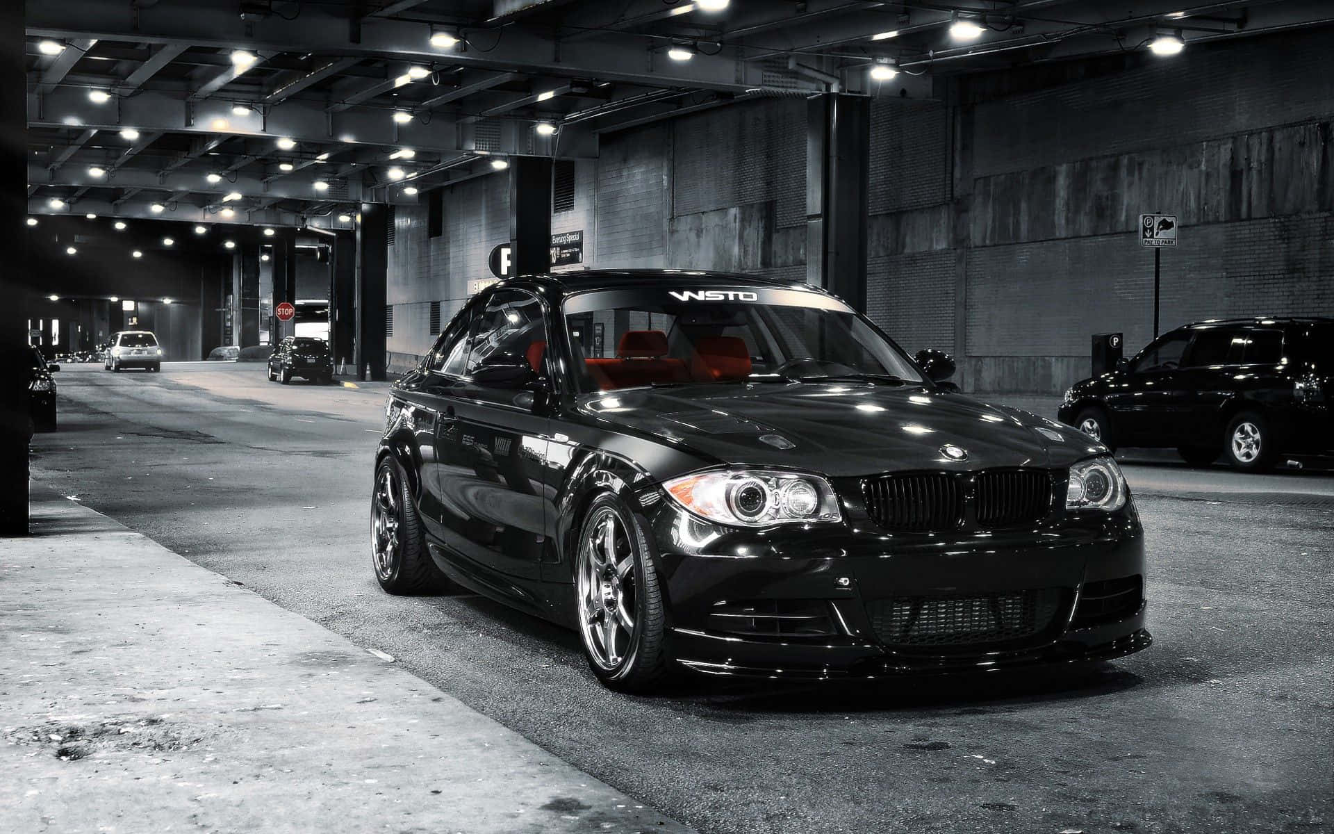 Bmw 1 Series 桌布 壁紙 — 一輛時尚的黑色BMW雙門轎跑車，紅色內裝，停在光線昏暗的地下停車場，有混凝土牆和頭頂燈光。