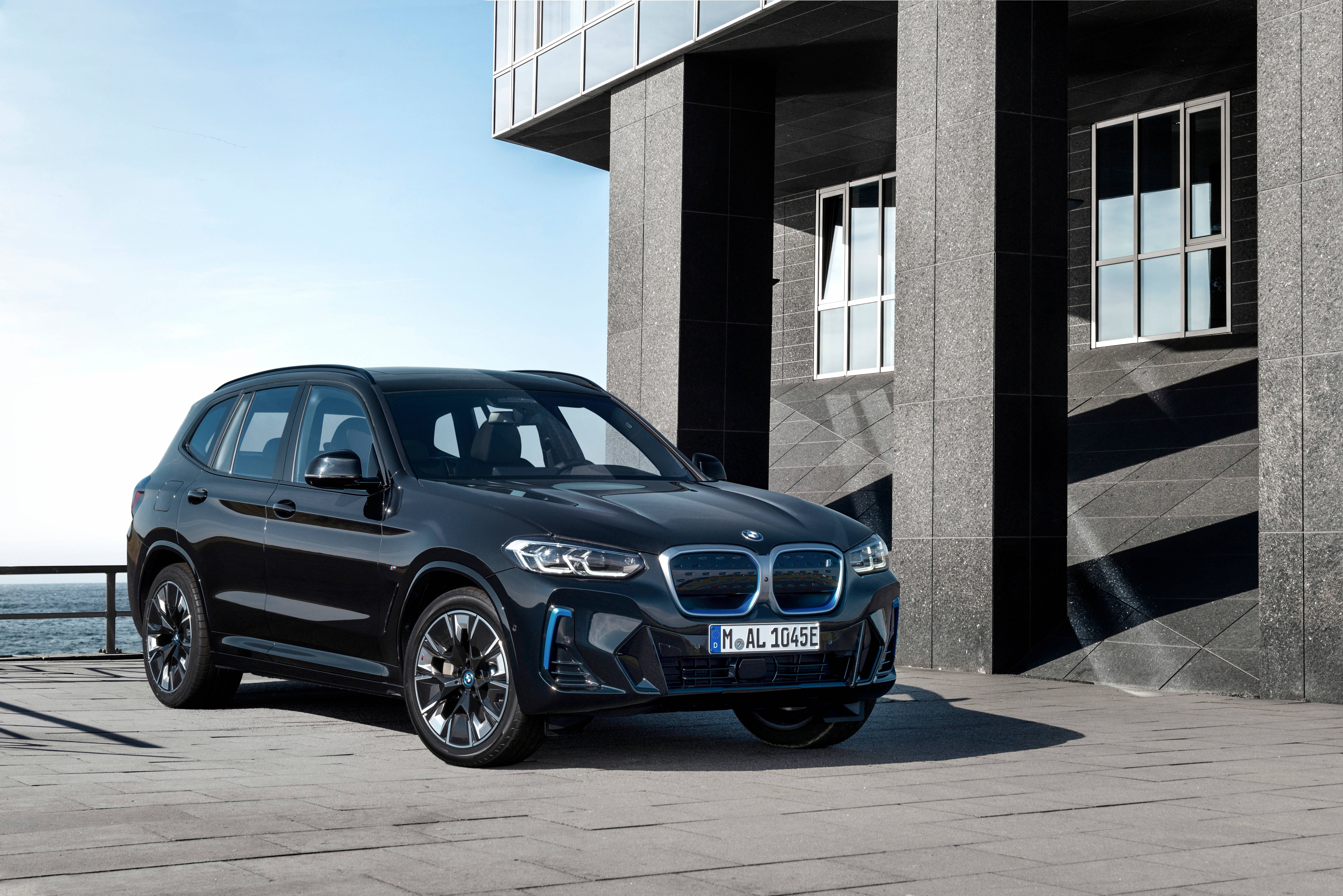 Black Bmw 桌布 壁紙 — 一輛時尚的黑色BMW電動SUV停在一棟現代混凝土建築旁，享有晴朗藍天下的海景。