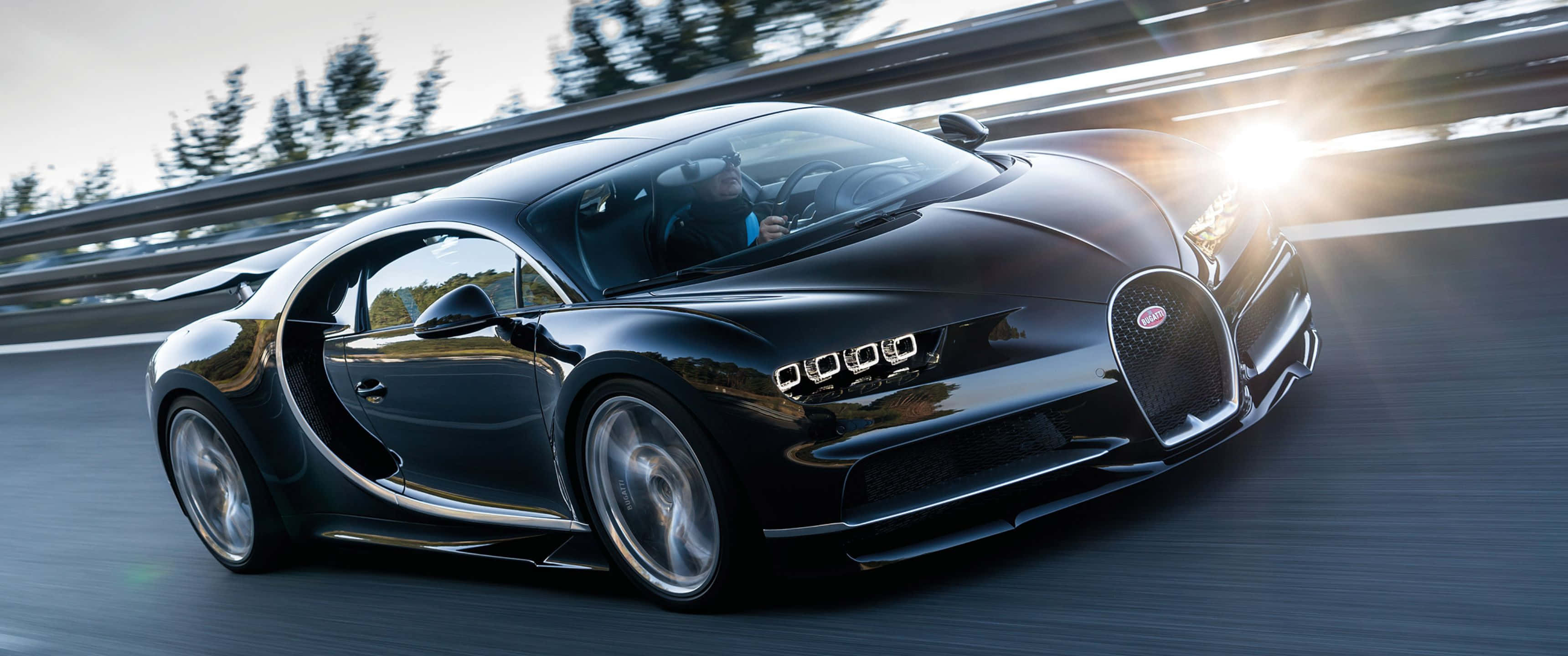 3440x1440p Bugatti 桌布 壁紙 — 一輛時尚的黑色Bugatti Chiron在蜿蜒的道路上行駛，背景有樹木和明亮的陽光。