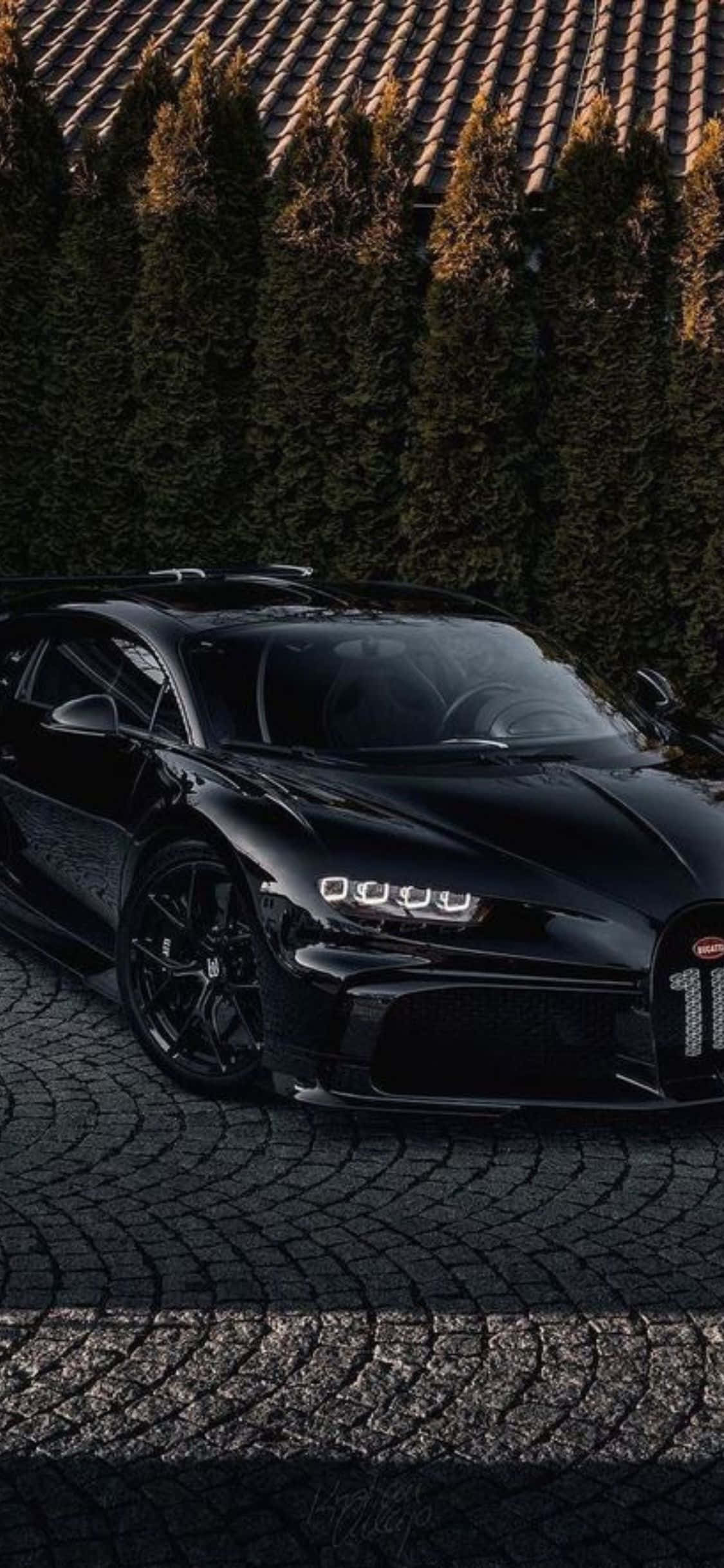 iPhone Xs Bugatti 桌布 壁紙 — 一輛時尚的黑色Bugatti Chiron停泊在鵝卵石車道上，背後有高大的常綠樹，沐浴在溫暖的金色光線中。