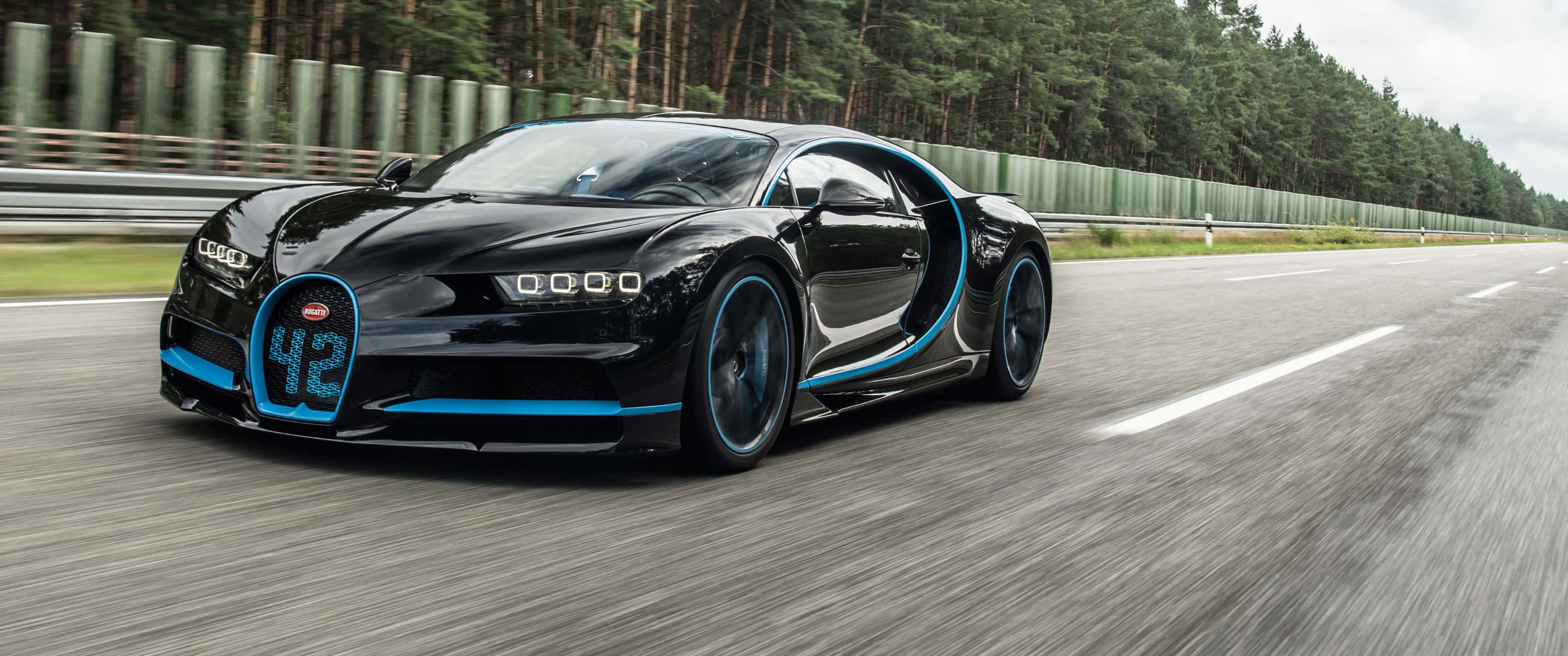 3440x1440p Bugatti 桌布 壁紙 — 一輛時尚的黑色Bugatti Chiron帶有藍色裝飾，在陰天之下沿著森林環繞的賽道飛馳。