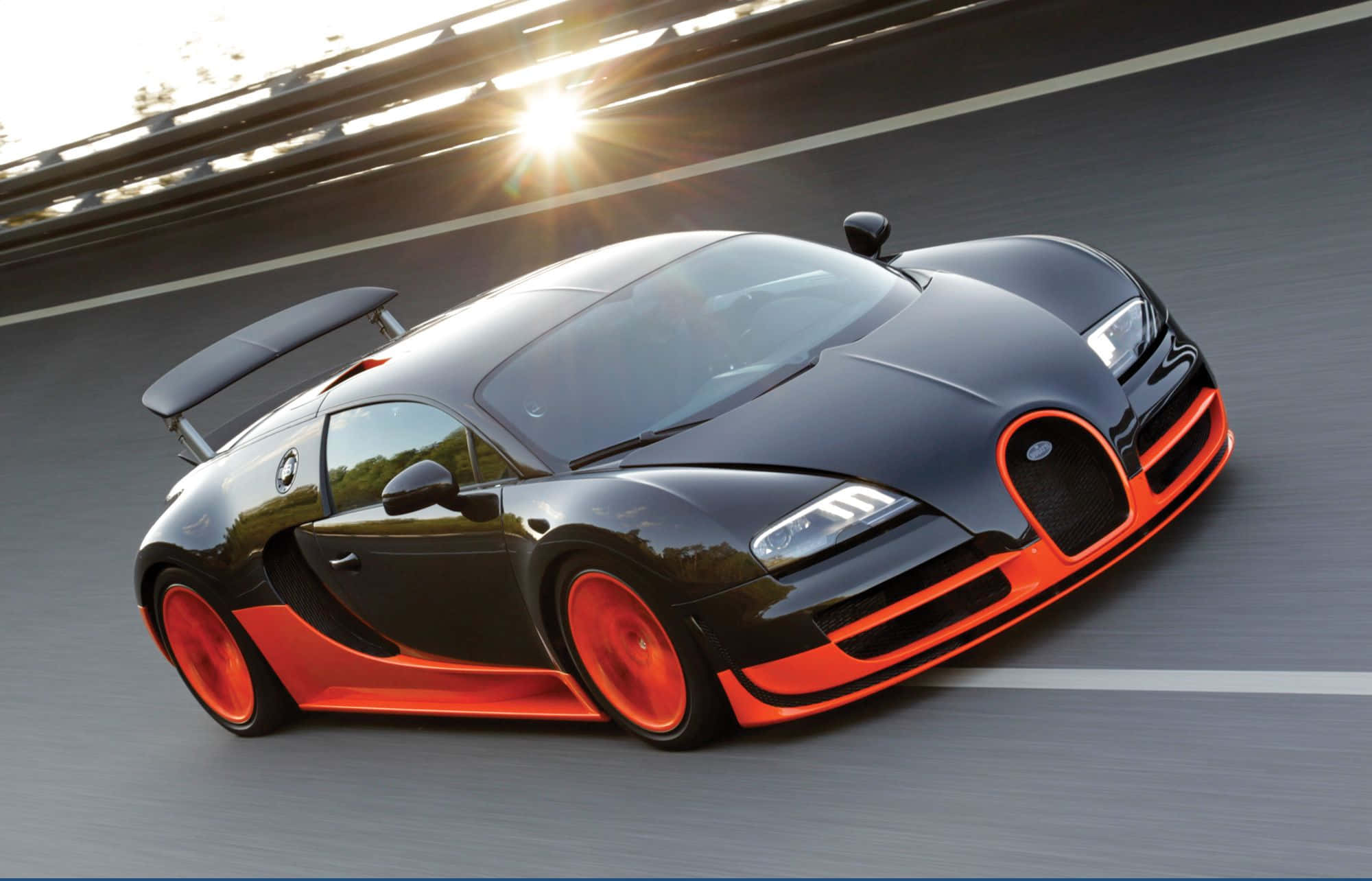 Best Bugatti 桌布 壁紙 — 一輛時尚的黑色Bugatti Veyron帶有鮮豔的橙色點綴，在陽光明媚的賽道上疾馳，其空氣動力學外形在強烈光芒下閃閃發光。