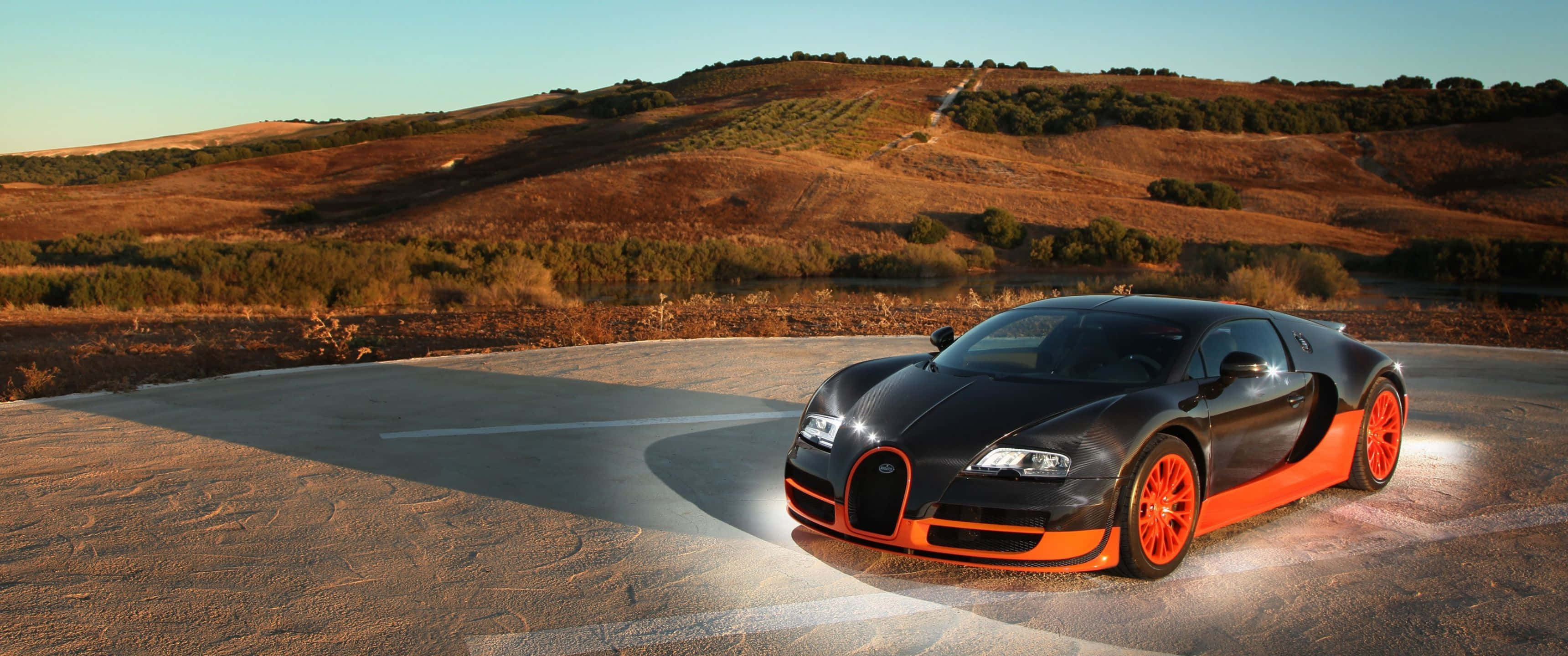 3440x1440p Bugatti 桌布 壁紙 — 一輛時尚的黑色Bugatti帶有鮮豔的橙色點綴，停在一條彎曲的道路上，背景是沐浴在溫暖夕陽光中的金色山丘。