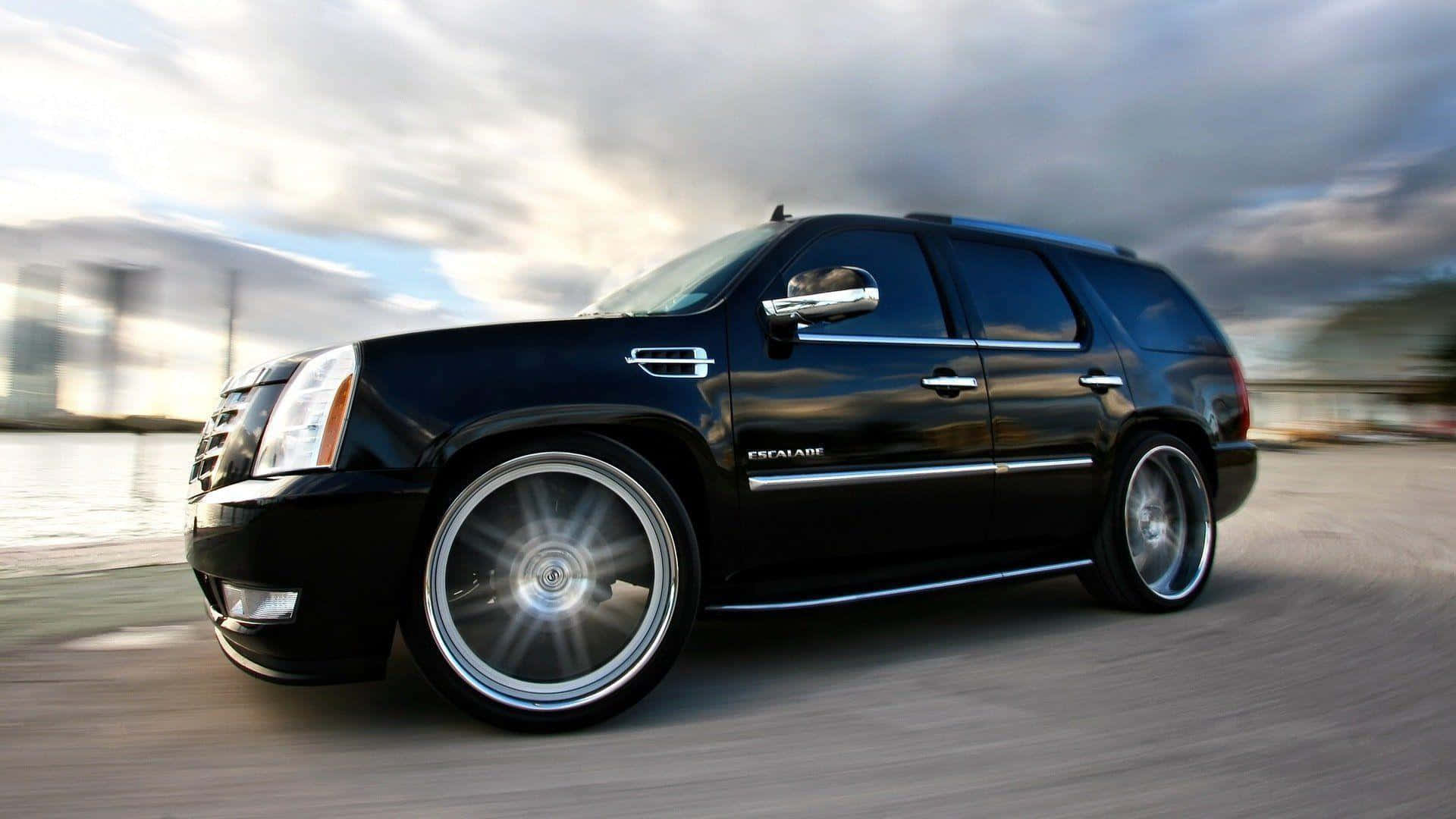 凱迪拉克 桌布 壁紙 — 一輛時尚的黑色Cadillac Escalade SUV配備定制鍍鉻輪圈，在陰天天空下沿著模糊的海濱道路飛馳。