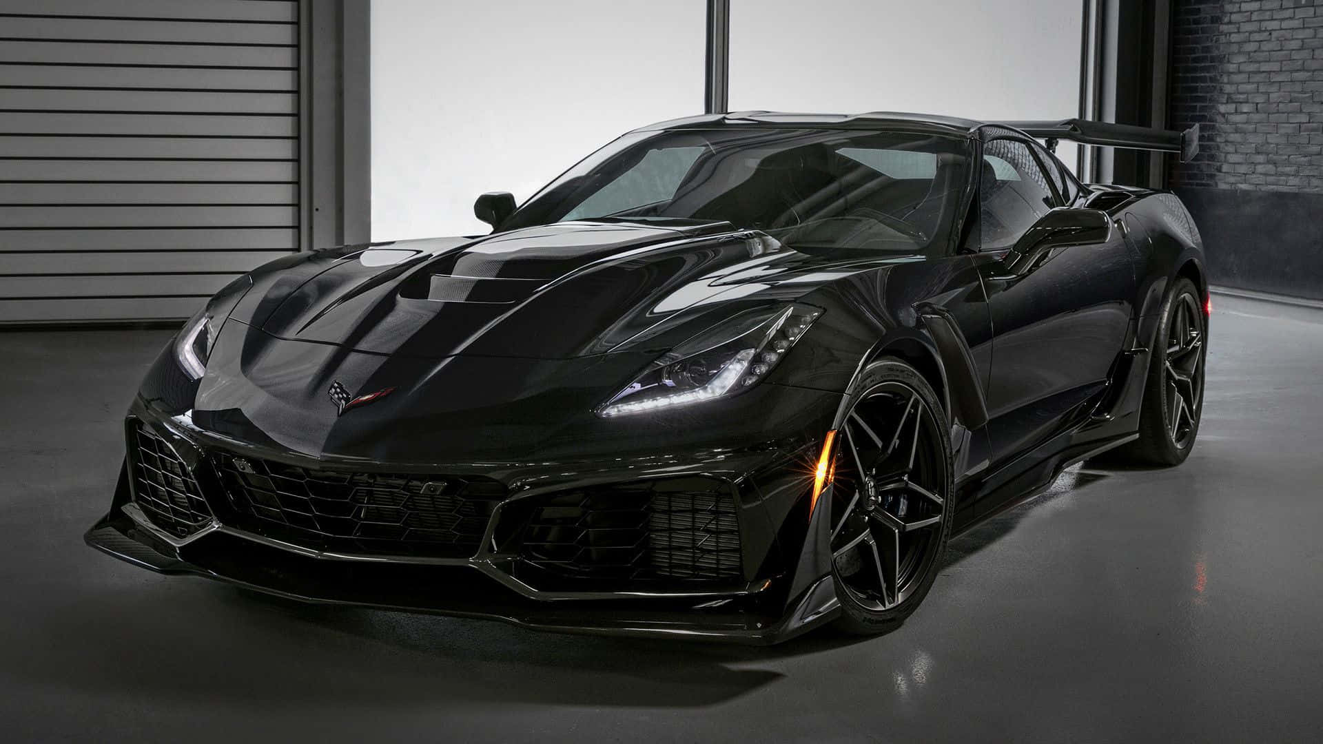 corvette 桌布 壁紙 — 一輛時尚的黑色Corvette跑車，配備發光頭燈和後擾流板，停放在設有磚牆和金屬門的現代化車庫中。