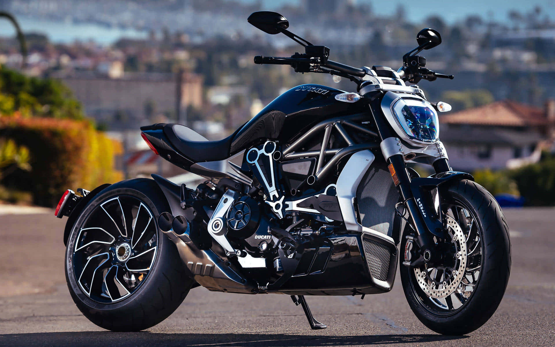 機車 桌布 壁紙 — 一輛時尚的黑色Ducati摩托車，帶有銀色點綴，停泊在陽光明媚的道路上，俯瞰著朦朧的海岸小鎮。