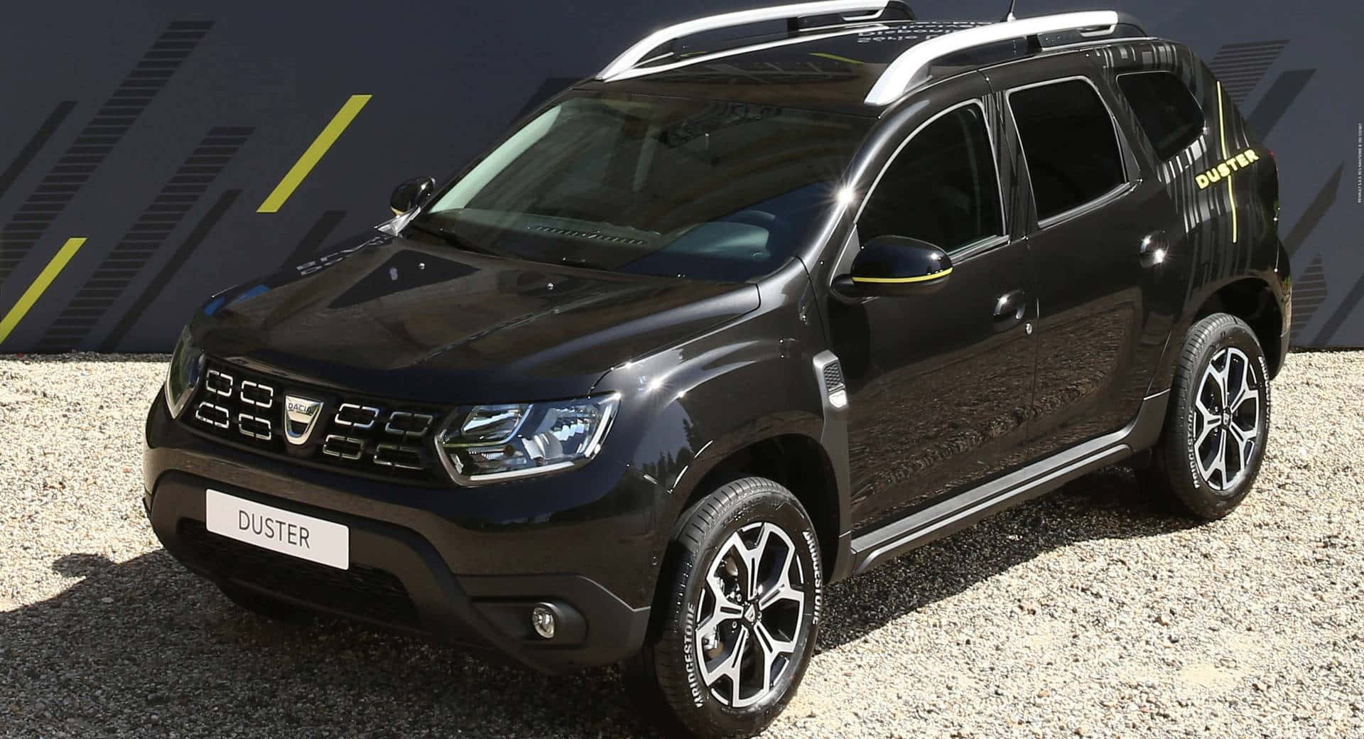 Dacia 桌布 壁紙 — 一輛時尚的黑色Duster SUV帶有黃色點綴，在明亮的日光下停在碎石上，背景是幾何圖案的深色牆壁。