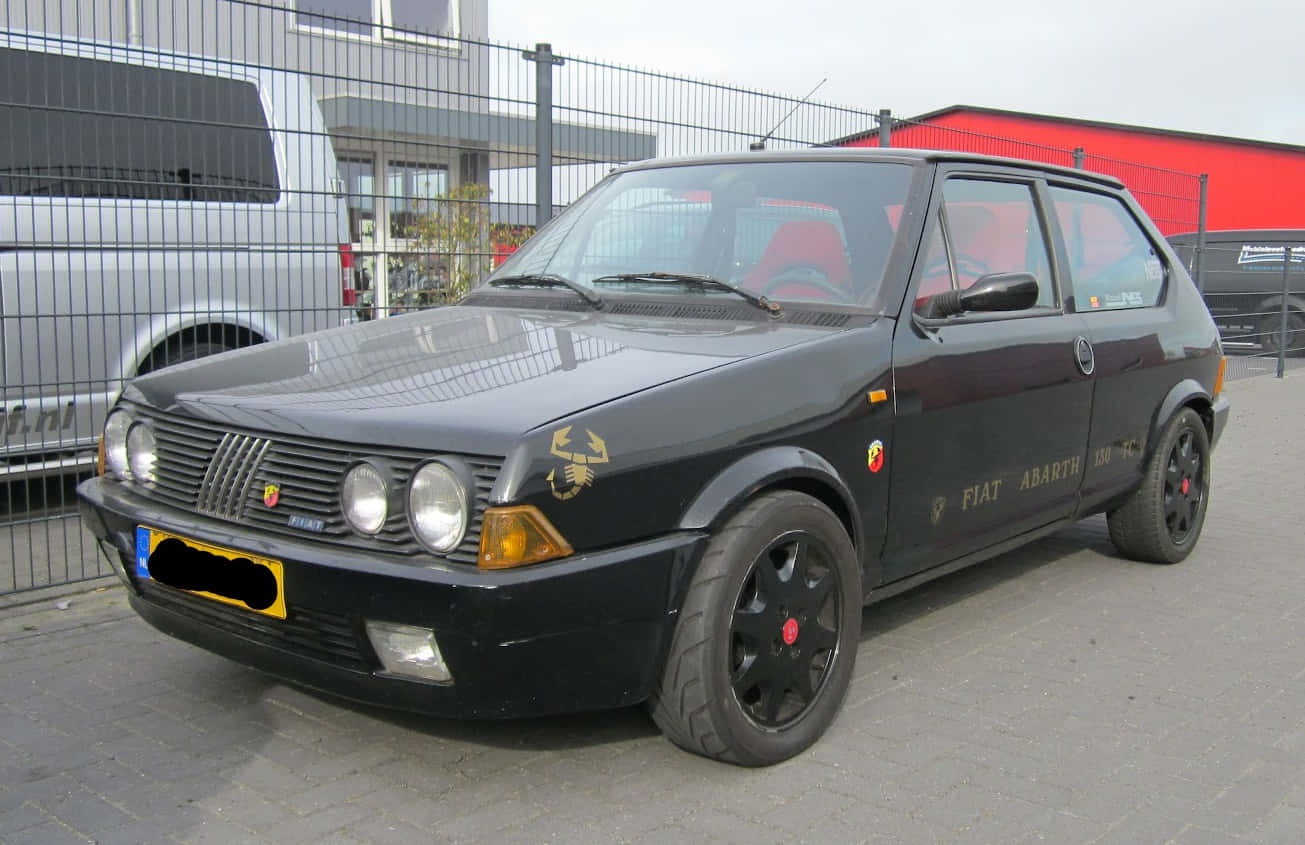 Fiat Ritmo 桌布 壁紙 — 一輛時尚的黑色Fiat Abarth 130 TC跑車停在人行道上，配備紅色內飾和金色蠍子標誌。