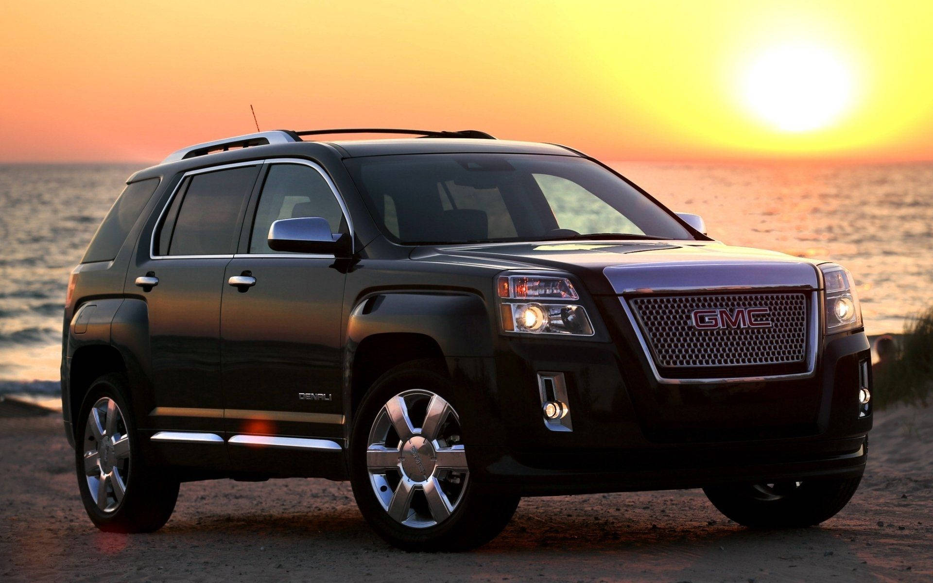 Gmc 桌布 壁紙 — 一輛時尚的黑色GMC Acadia SUV停在日落時分的沙灘上，金色光線反射在它的鉻合金輪圈和格柵上。