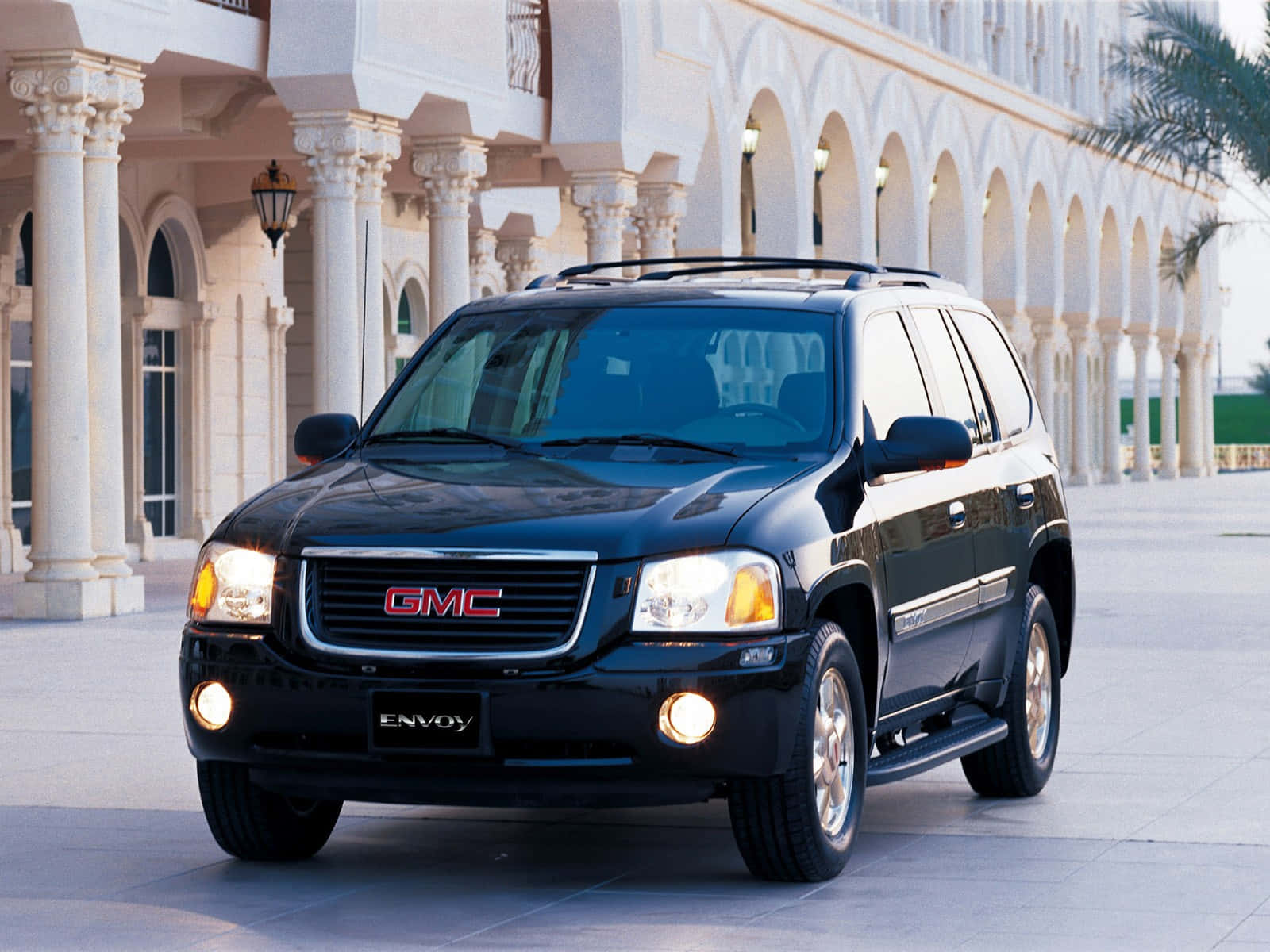Gmc Envoy 桌布 壁紙 — 一輛時尚的黑色GMC Envoy SUV在日光下閃耀，停在一座帶拱形步道的宏偉白色柱廊建築前。