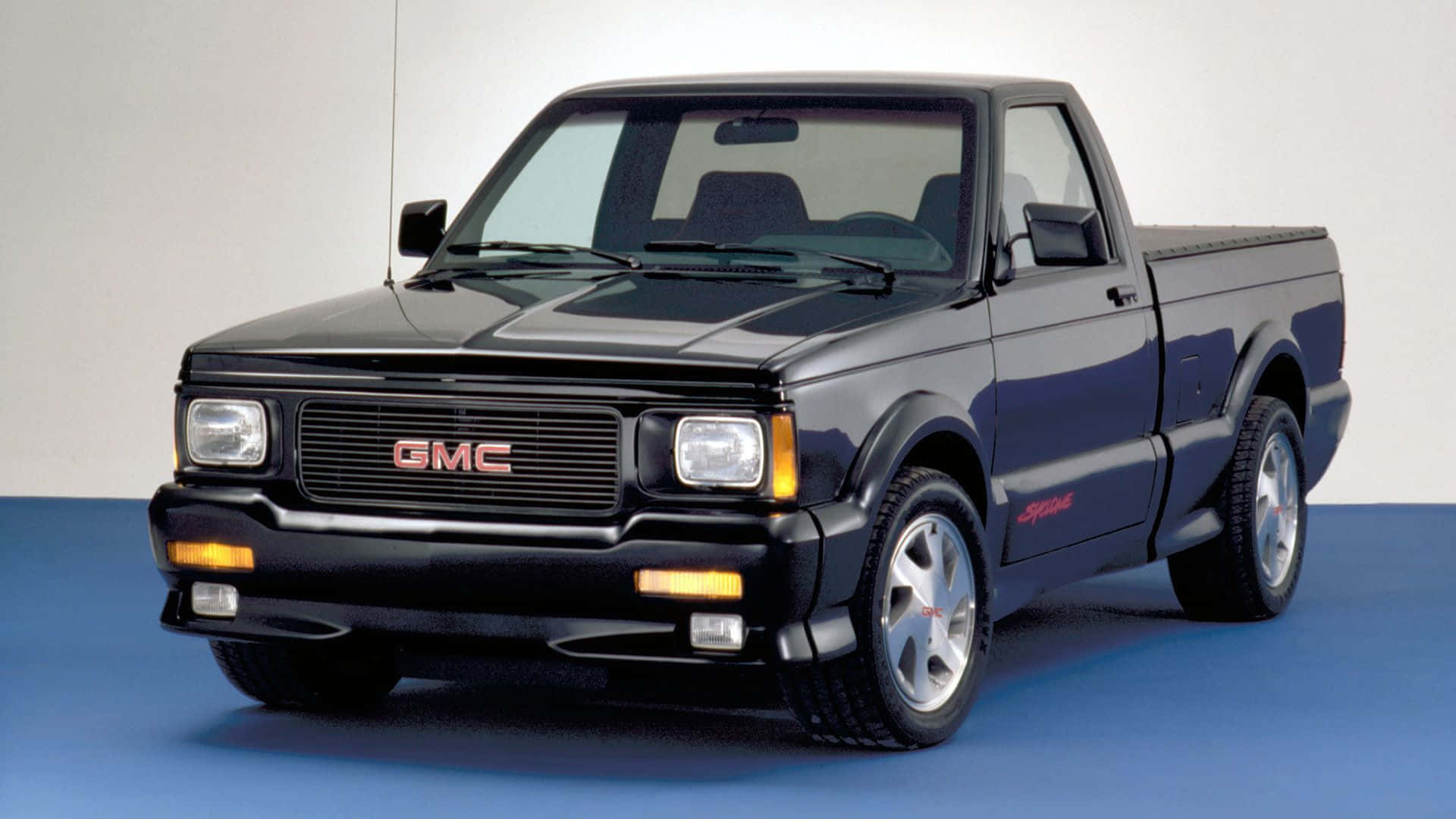 Gmc Syclone 桌布 壁紙 — 一輛時尚的黑色GMC Syclone皮卡車，帶有紅色點綴和銀色輪圈，擺放在藍色地板和白色牆壁前。