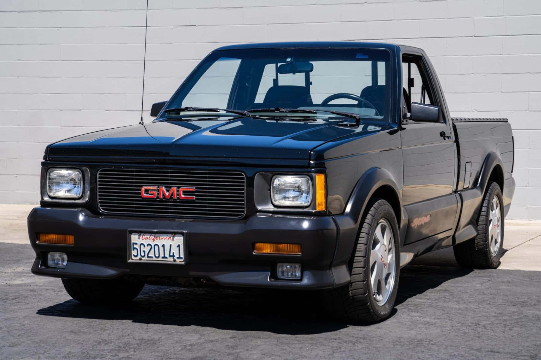 Gmc Syclone 桌布 壁紙 — 一輛時尚的黑色GMC Syclone皮卡在明亮的陽光下閃耀，背景是潔淨的白色磚牆。