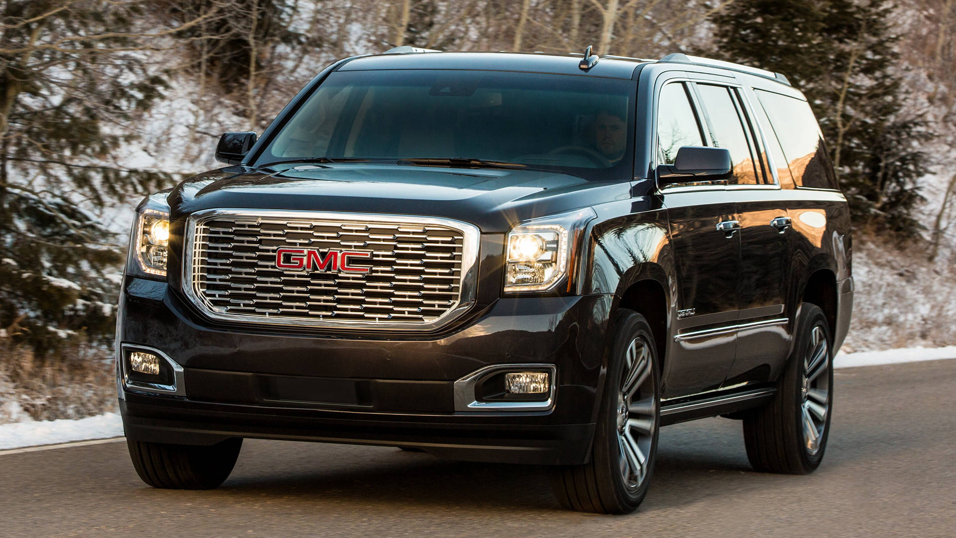 Gmc 桌布 壁紙 — 一輛時尚的黑色GMC Yukon SUV在柔和的日光下行駛在一條被冬日禿樹環繞的雪路上。