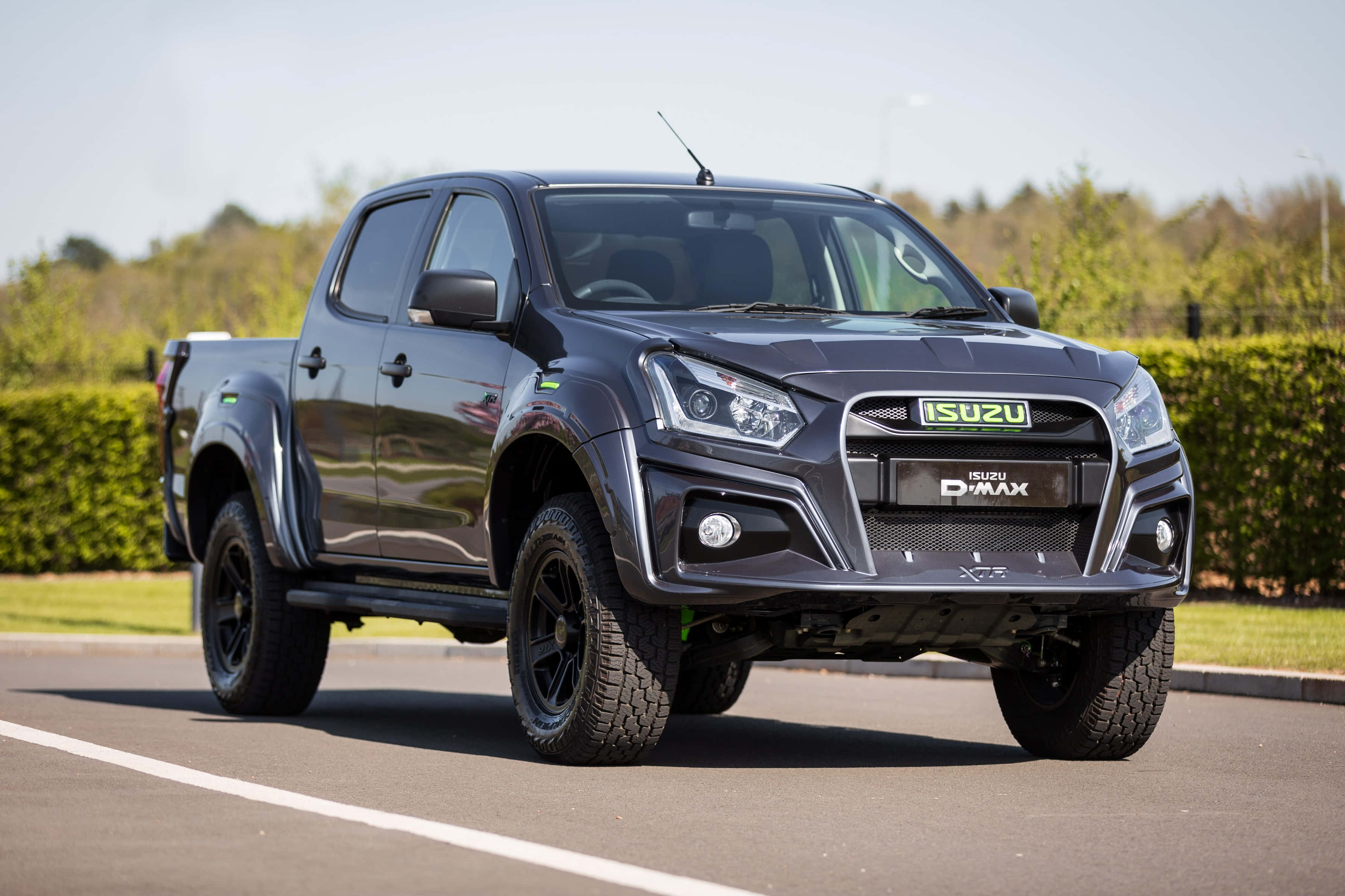 Isuzu 桌布 壁紙 — 一輛時尚的黑色Isuzu D-MAX皮卡車停在瀝青路上，背景有綠色籬笆和樹木，天空晴朗。