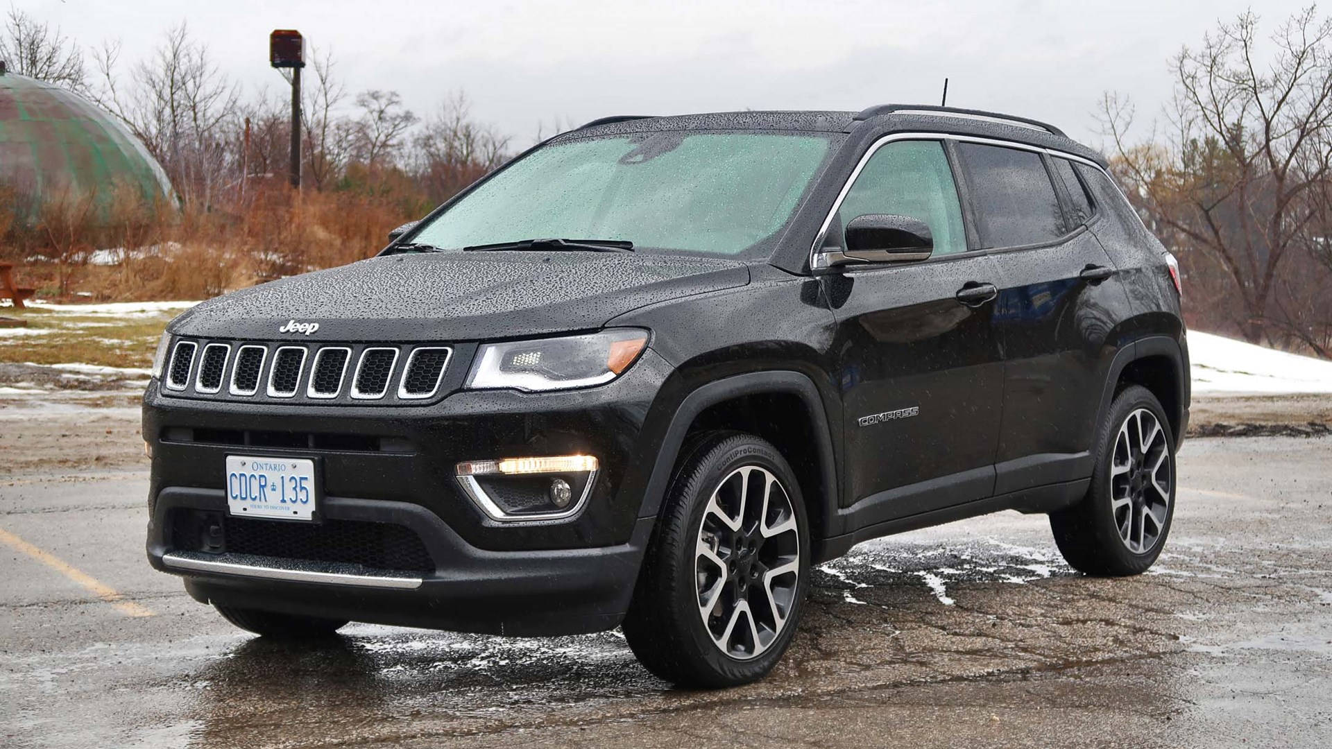 Road 桌布 壁紙 — 一輛時尚的黑色Jeep Compass停放在濕漉漉的瀝青場地上，背景有雪堆和光禿禿的樹木，天空多雲。