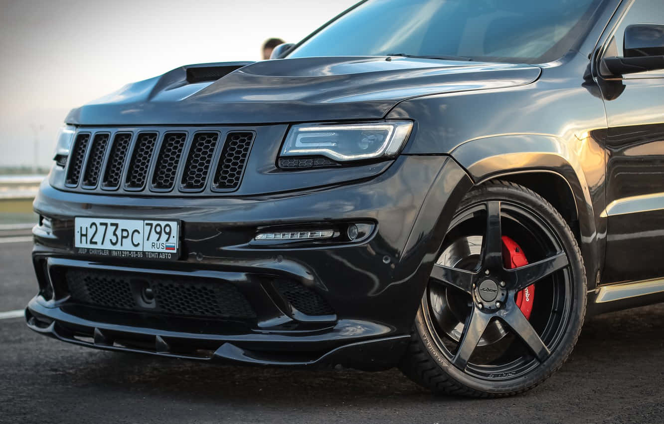 Jeep Trackhawk 桌布 壁紙 — 一輛時尚的黑色Jeep Grand Cherokee Trackhawk，配備紅色制動卡鉗和定制輪圈，停在明亮的日光下的瀝青路面上。