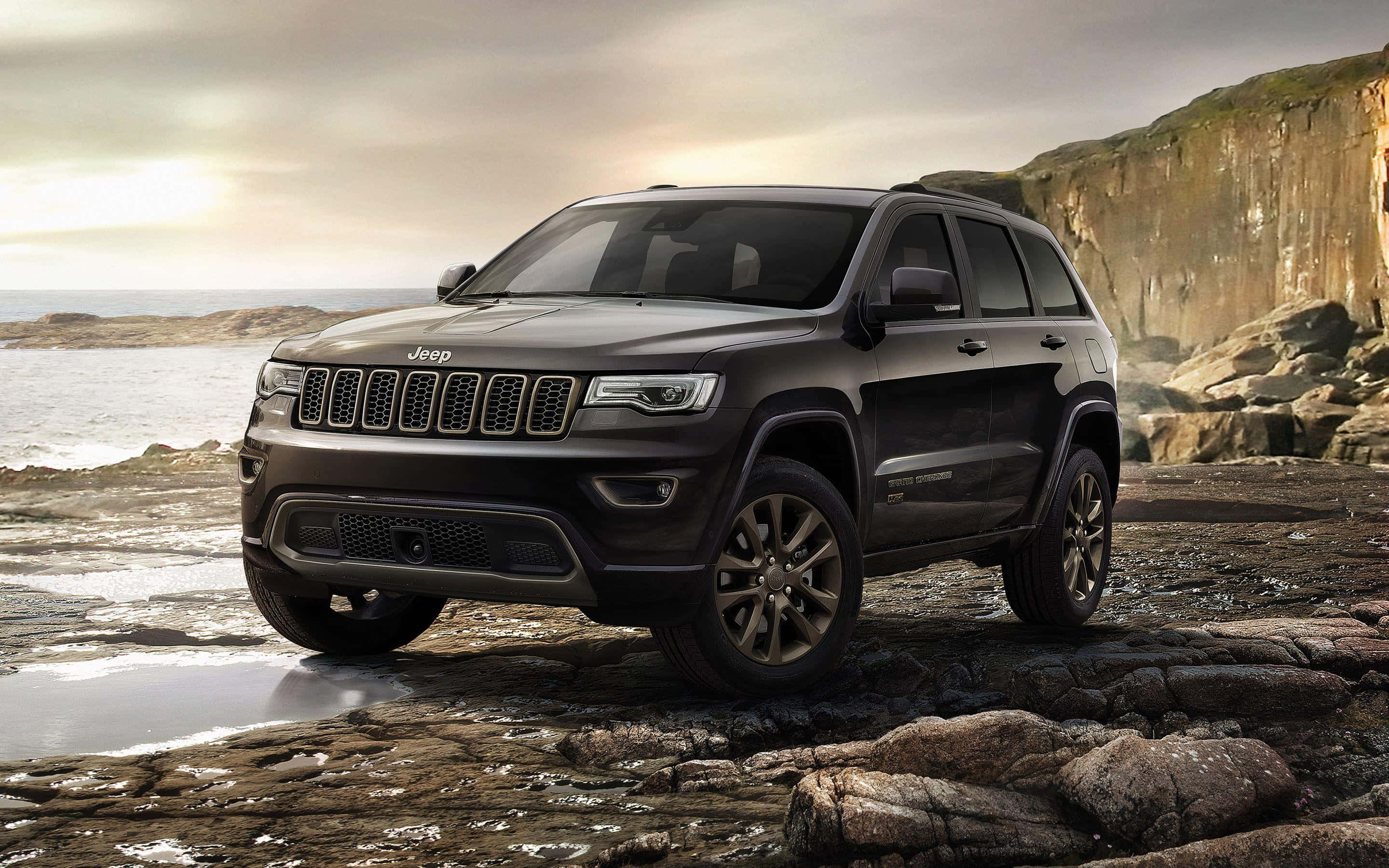 Jeep Wagoneer 桌布 壁紙 — 一輛時尚的黑色Jeep Grand Cherokee停在濕漉漉的海岸岩石上，背景是戲劇性的懸崖和海洋，天空多雲。