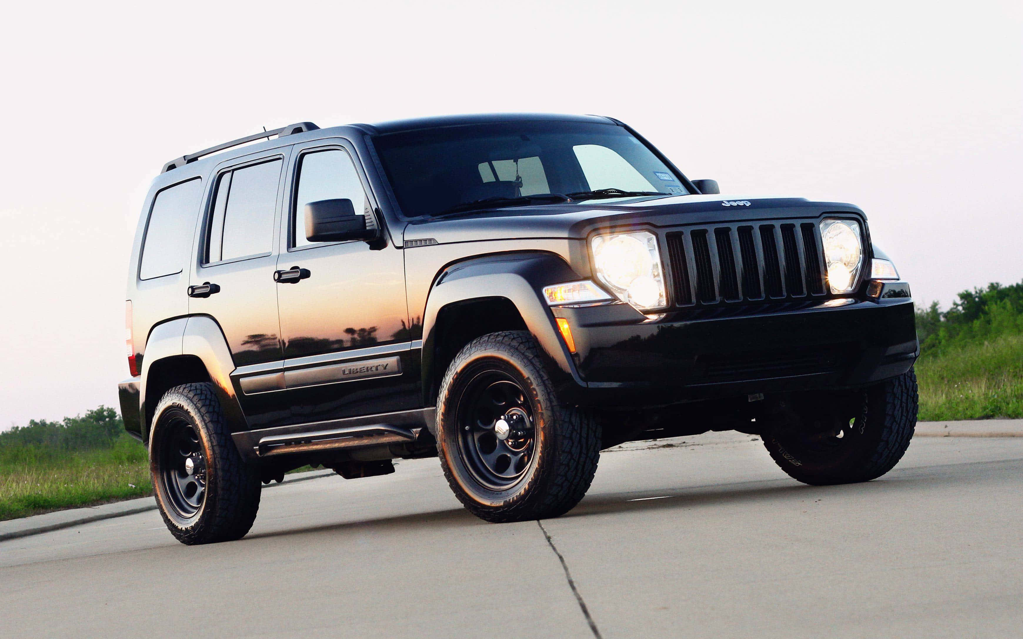 Jeep Liberty 桌布 壁紙 — 一輛時尚的黑色Jeep Liberty SUV，車頭燈閃爍，停在日落時分的柏油路上，背景是草地田野。