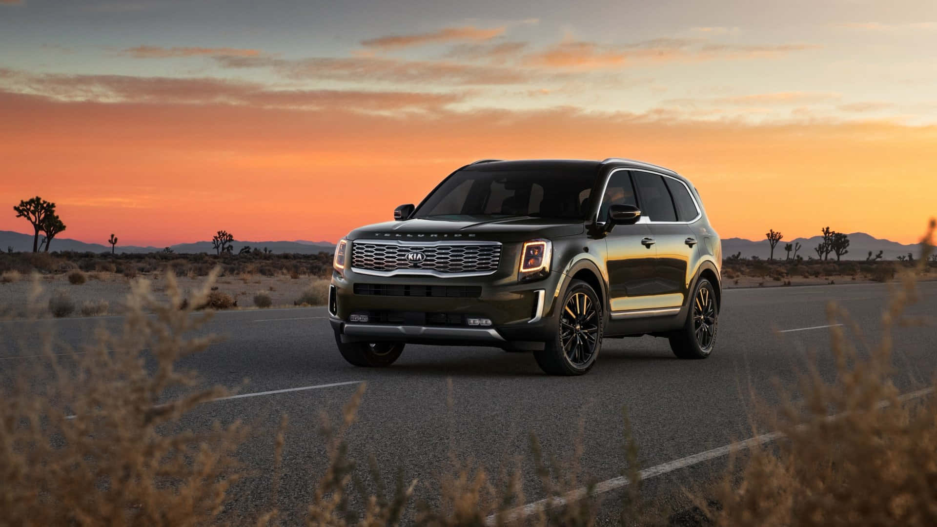 Kia Telluride 桌布 壁紙 — 一輛時尚的黑色Kia Telluride SUV在充滿活力的橙色和粉紅色日落天空下行駛在沙漠公路上。