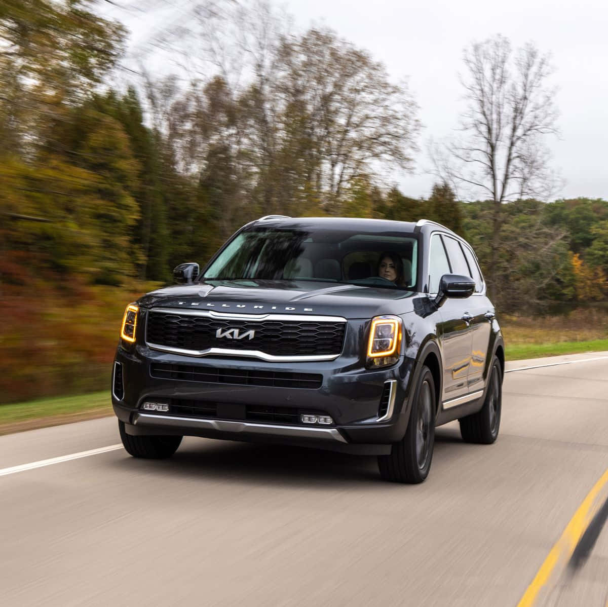 Kia Telluride 桌布 壁紙 — 一輛時尚的黑色Kia Telluride SUV在秋日樹林中沿著蜿蜒的道路行駛，樹葉模糊。