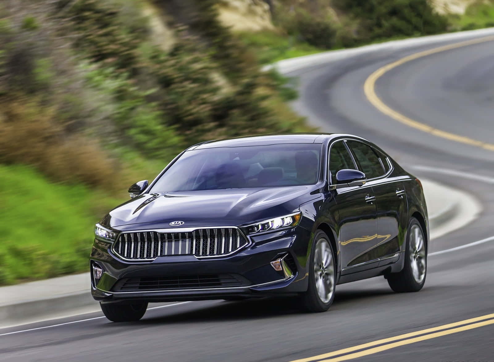 Kia Cadenza 桌布 壁紙 — 一輛時尚的黑色Kia轎車在蜿蜒的山路上疾馳，周圍是模糊的綠色樹葉和黃色車道標線。