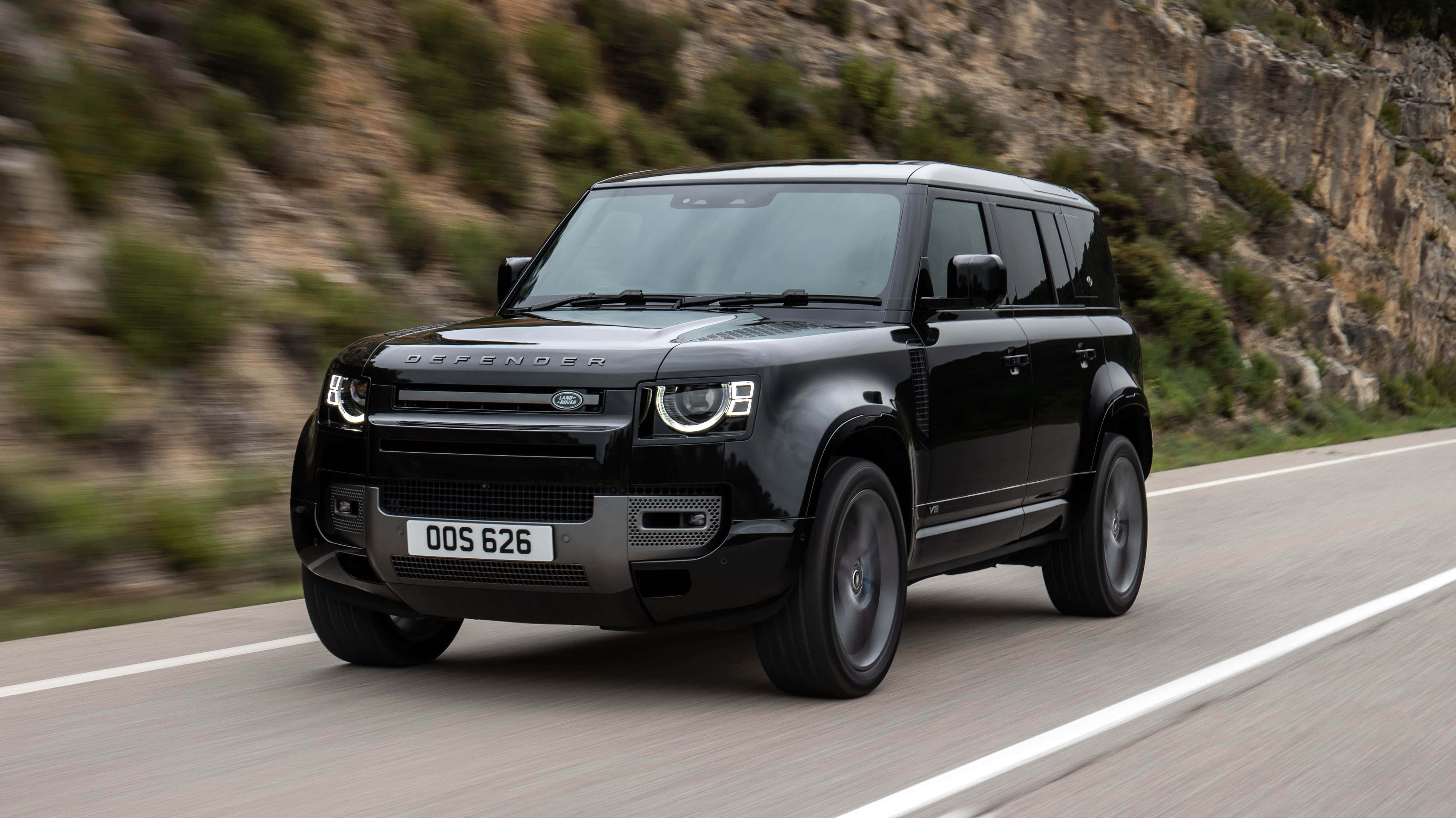 Land Rover Defender 桌布 壁紙 — 一輛時尚的黑色Land Rover Defender在蜿蜒的山路上疾馳，背景是模糊的綠意和岩石峭壁。