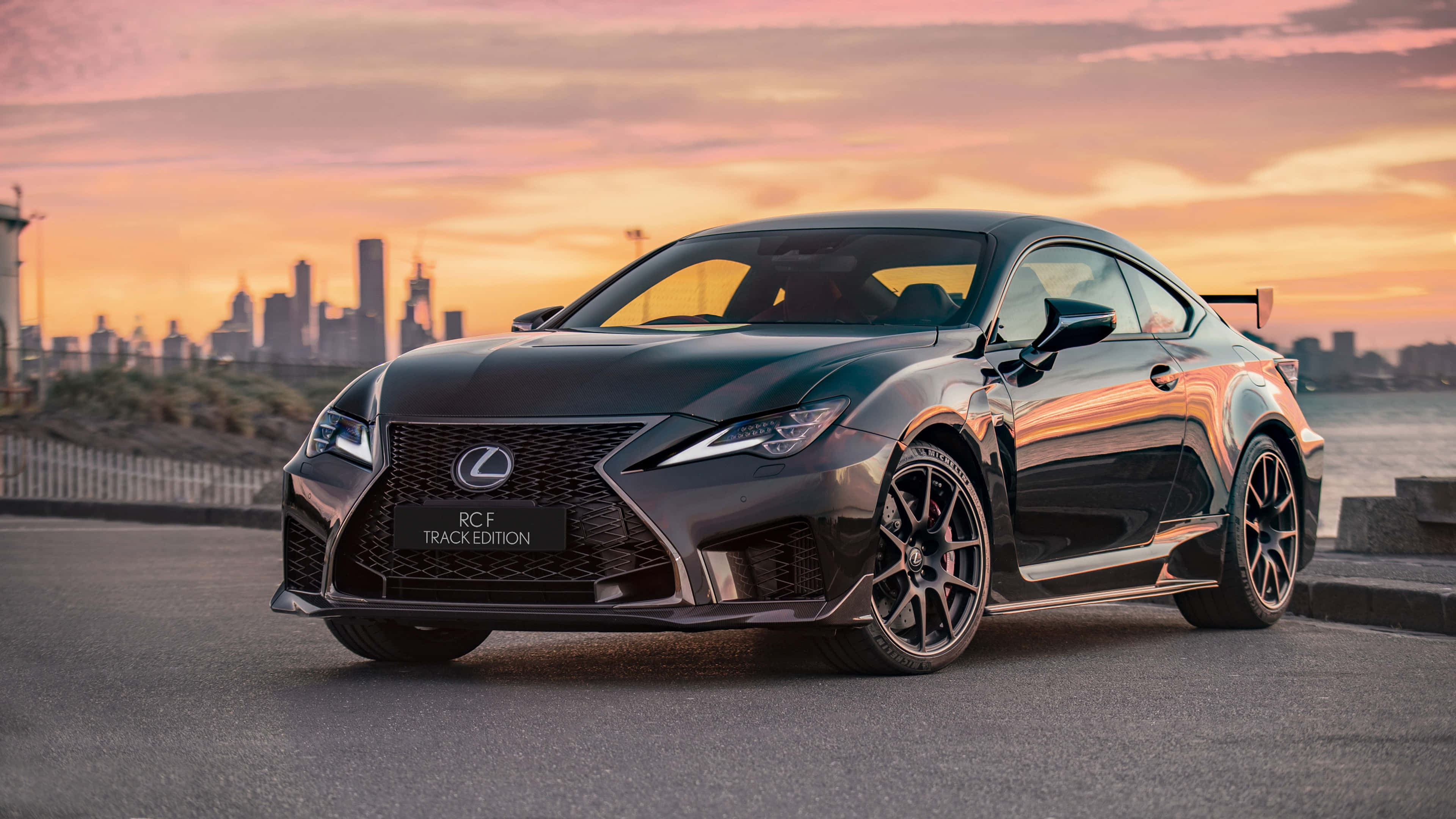 Rc Car 桌布 壁紙 — 一輛時尚的黑色Lexus RCF Track Edition在充滿活力的日落天空下閃耀，背景是城市天際線。