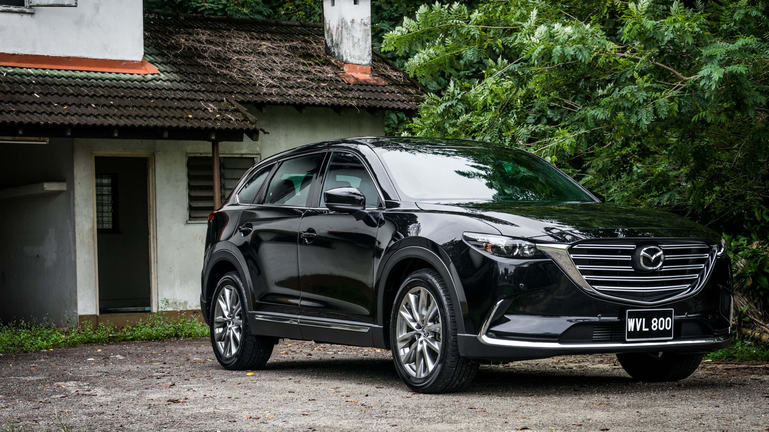 Mazda Cx-9 桌布 壁紙 — 一輛時尚的黑色Mazda CX-9 SUV停在碎石路上，前面是一間有茂密綠色植物的鄉村風格房屋。