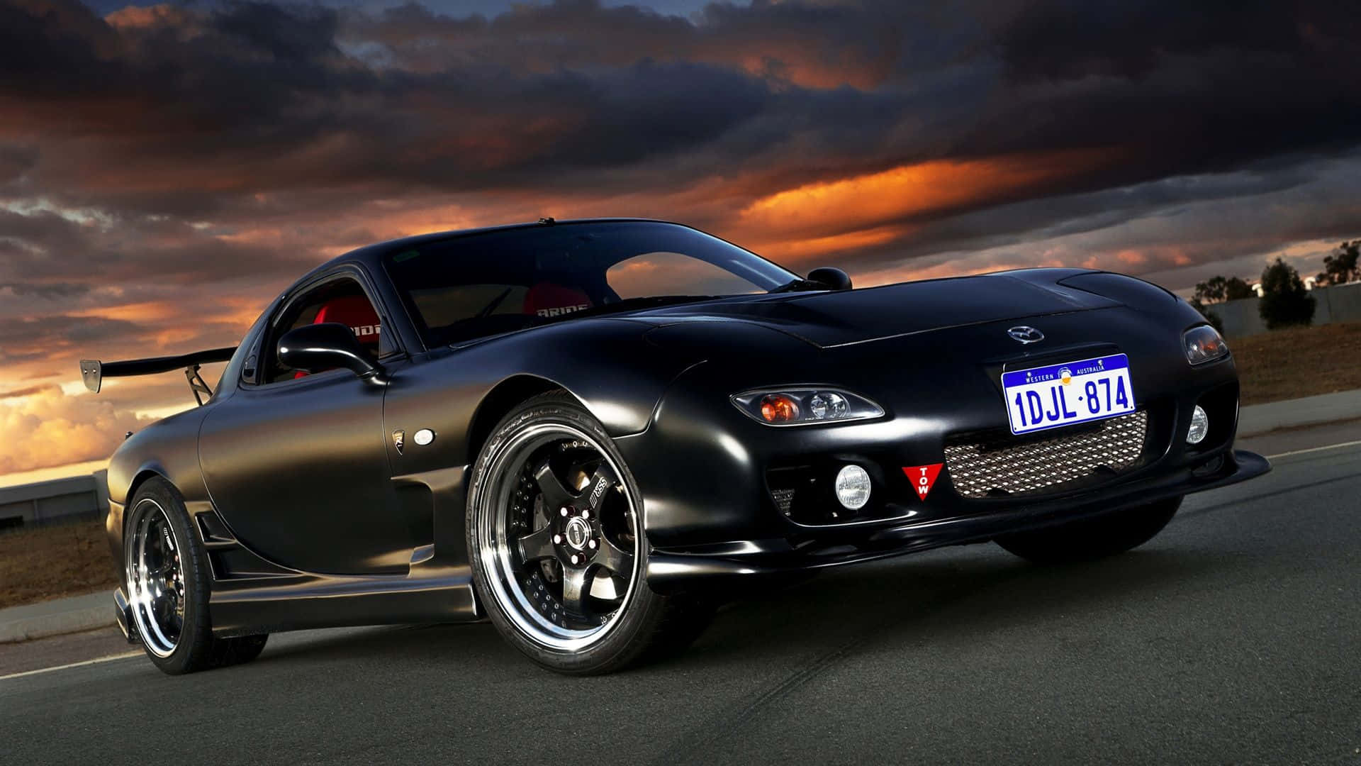暴風 桌布 壁紙 — 一輛時尚的黑色Mazda RX-7跑車，配備定制輪圈和後擾流板，在戲劇性的日落天空下行駛在道路上。