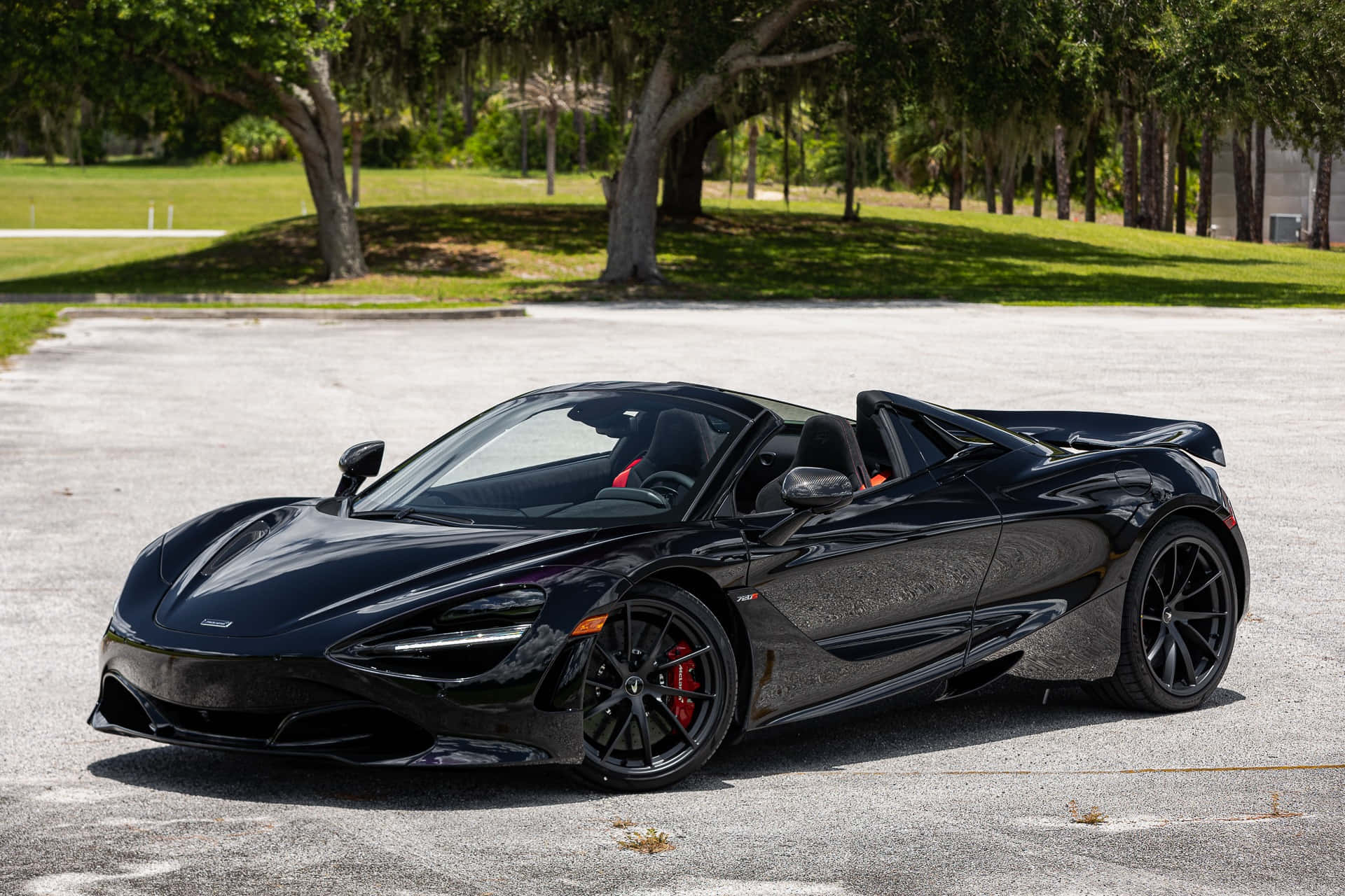 麥拉倫 桌布 壁紙 — 一輛時尚的黑色McLaren 720S Spider敞篷車停在柏油路上，陽光燦爛，背景是茂密的綠樹。