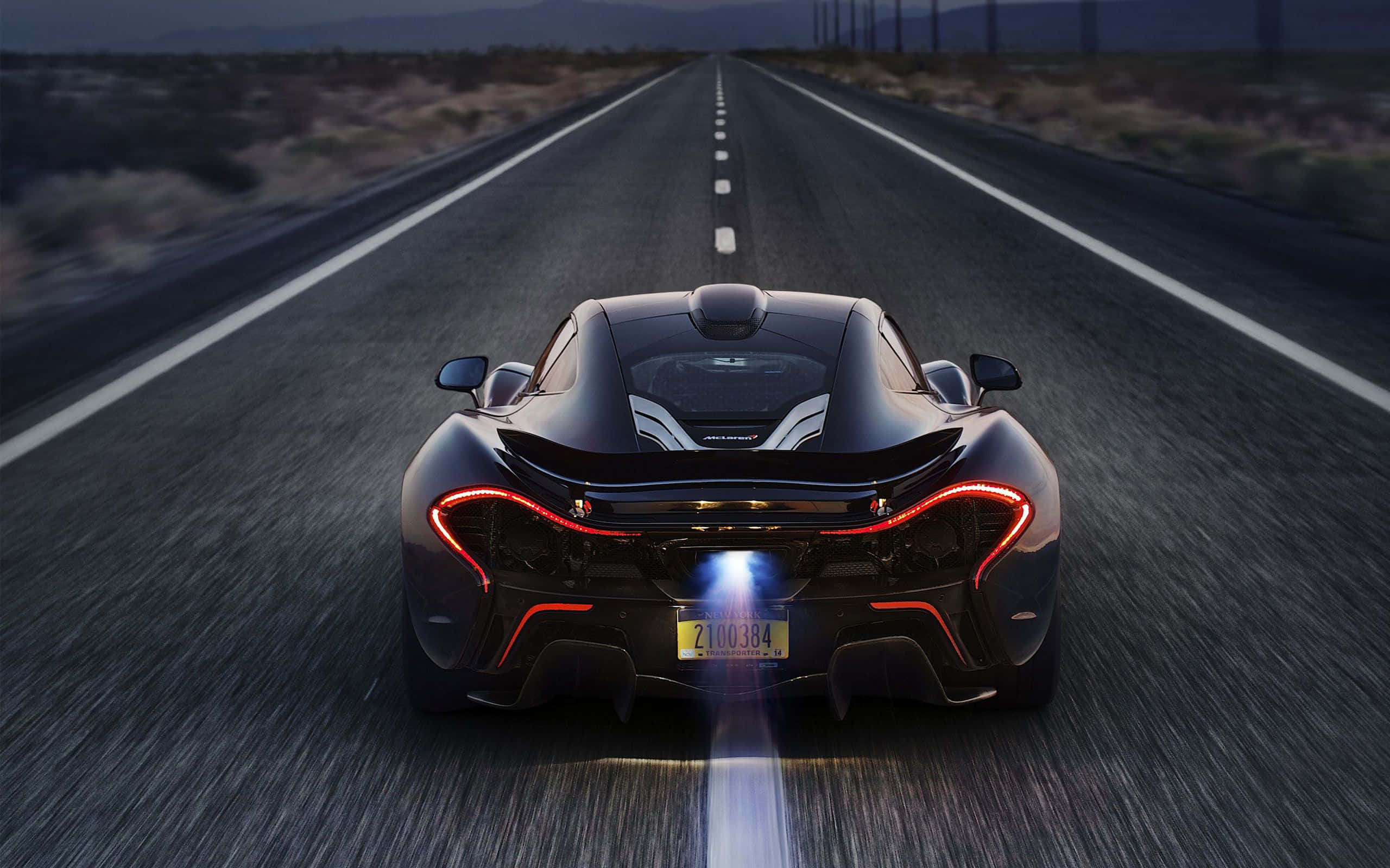 Cool Mclaren 桌布 壁紙 — 一輛時尚的黑色McLaren P1在黃昏時分在沙漠高速公路上飛馳，帶有發光的紅色尾燈和藍色排氣火焰。