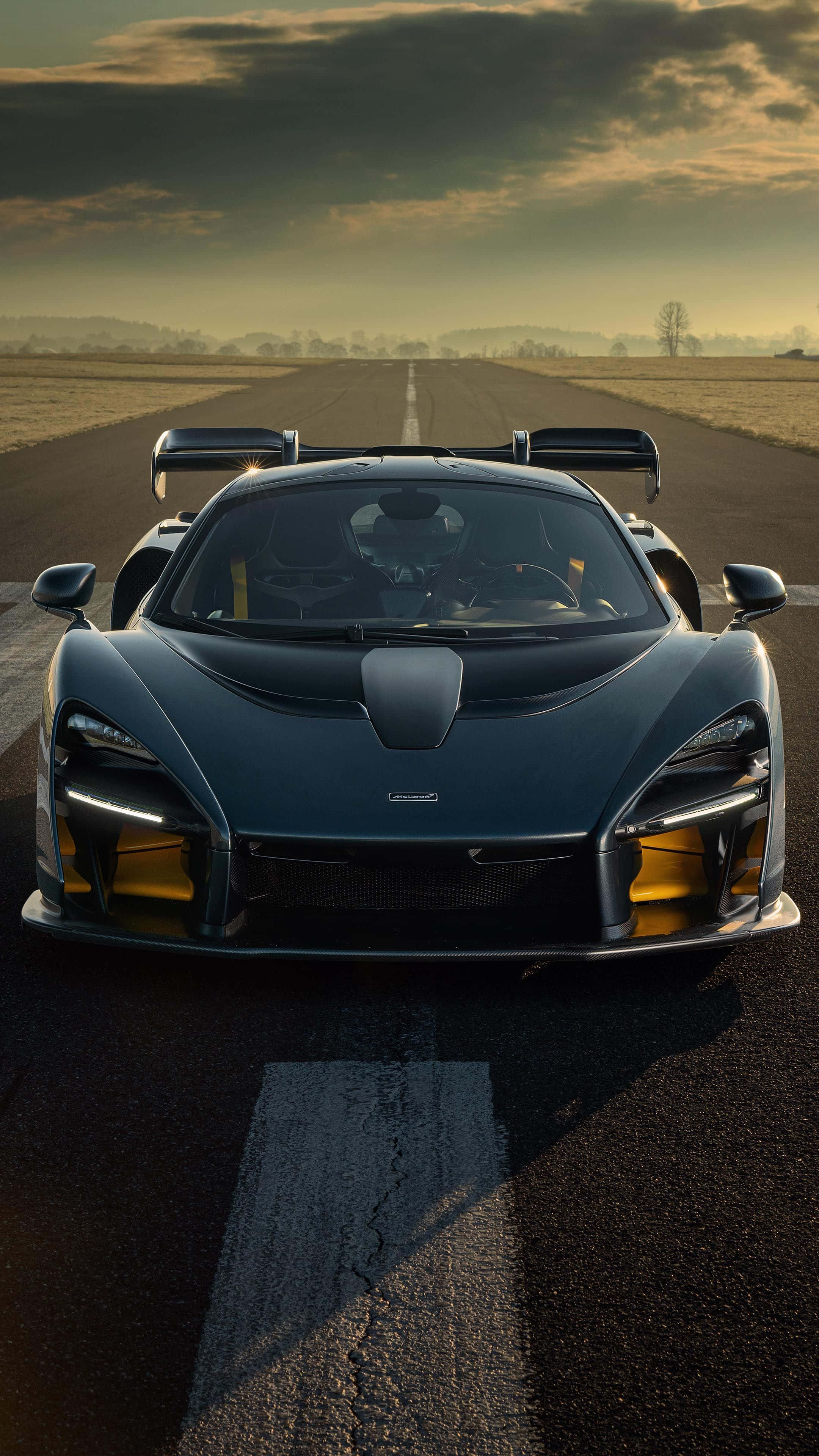 高清 桌布 壁紙 — 一輛時尚的黑色McLaren Senna跑車帶有發光的黃色點綴，在戲劇性的金色日落天空下，面向前方停在長長的瀝青跑道上。