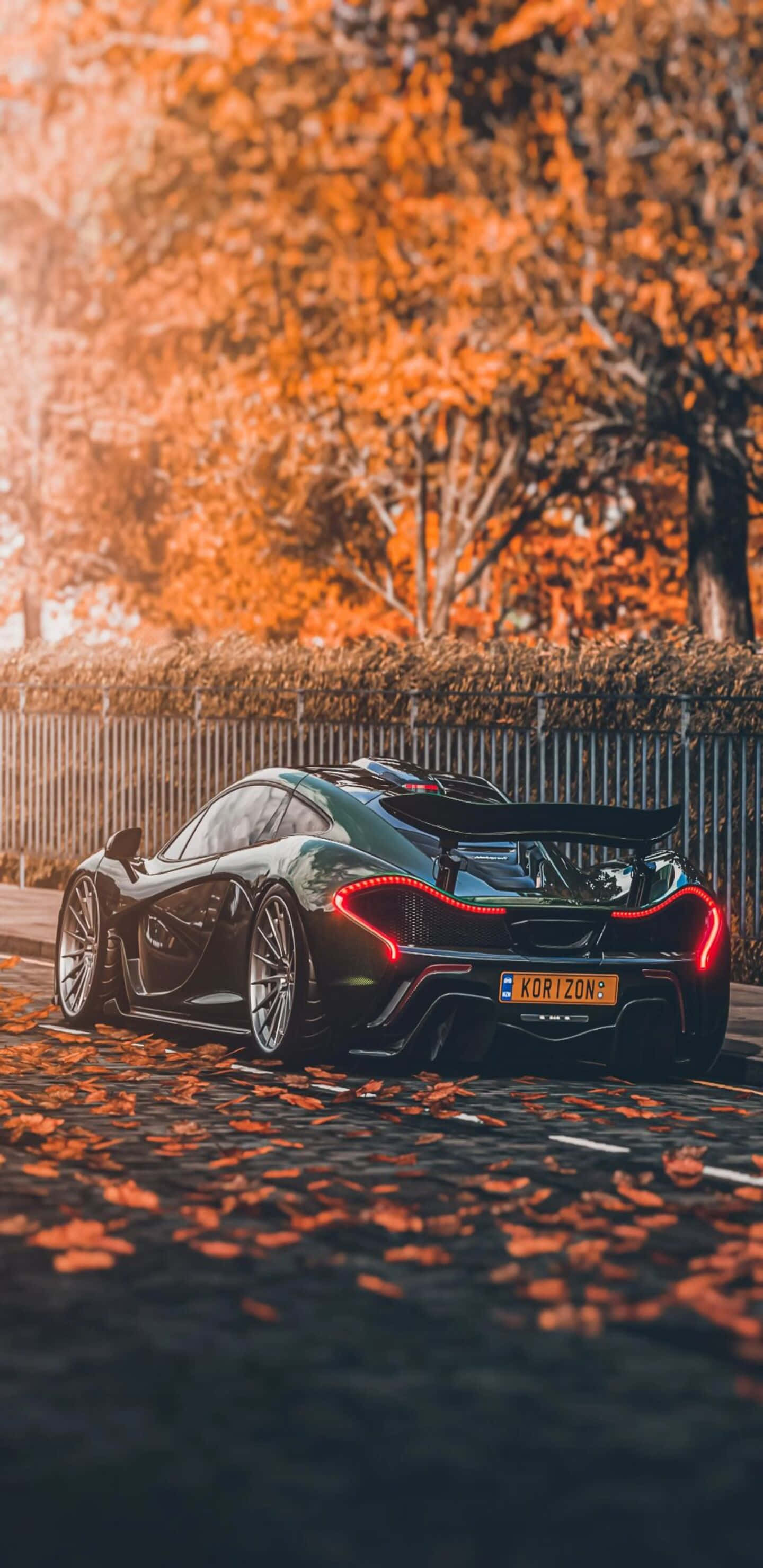 黑色 桌布 壁紙 — 一輛時尚的黑色McLaren跑車，帶有發光的紅色尾燈，停在鋪滿秋葉的鵝卵石街道上，沐浴在溫暖的金色陽光下。