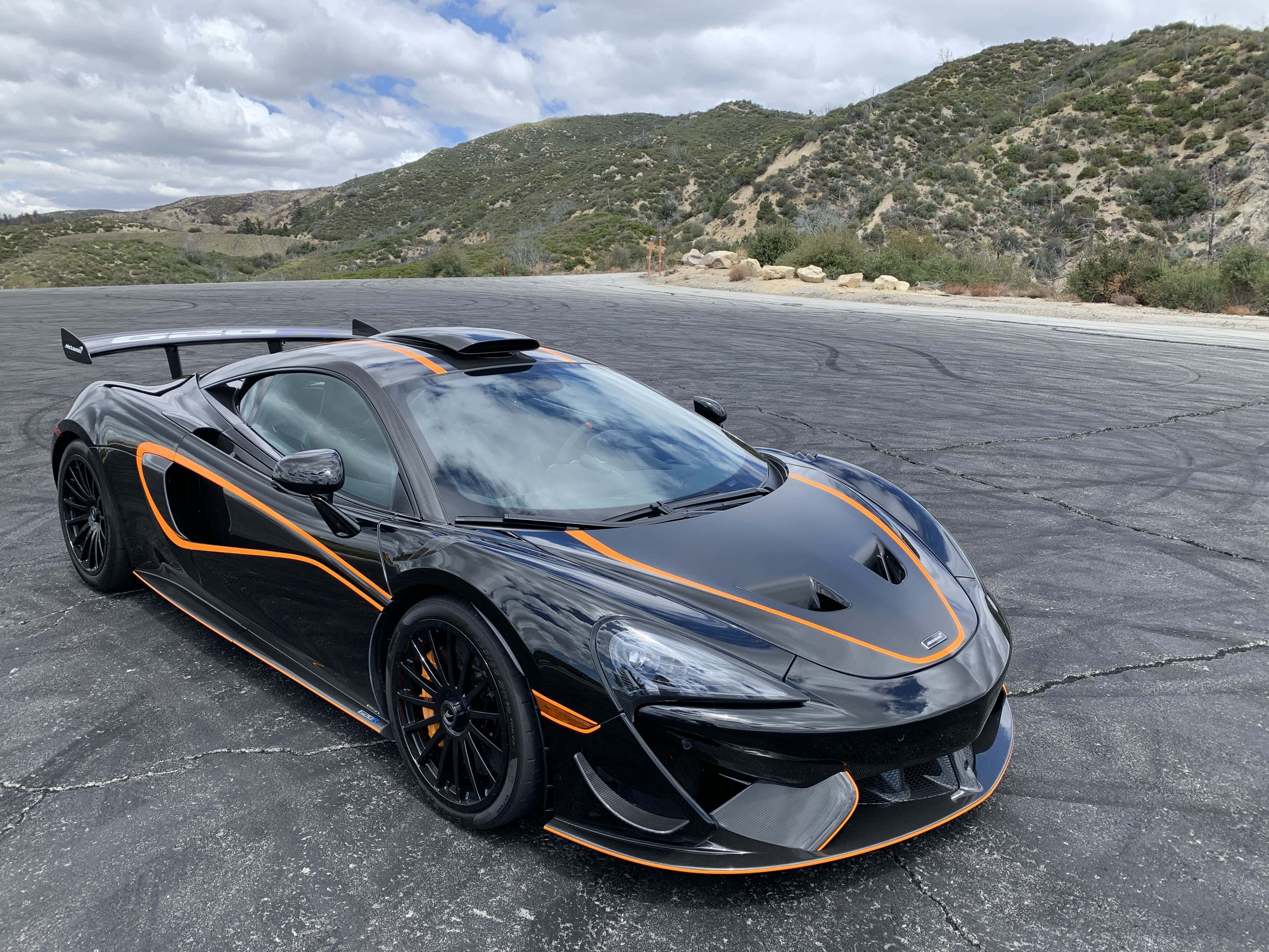麥拉倫 桌布 壁紙 — 一輛時尚的黑色McLaren跑車帶有橙色點綴，停在裂縫的瀝青地上，背景是連綿起伏的山丘和陰天。