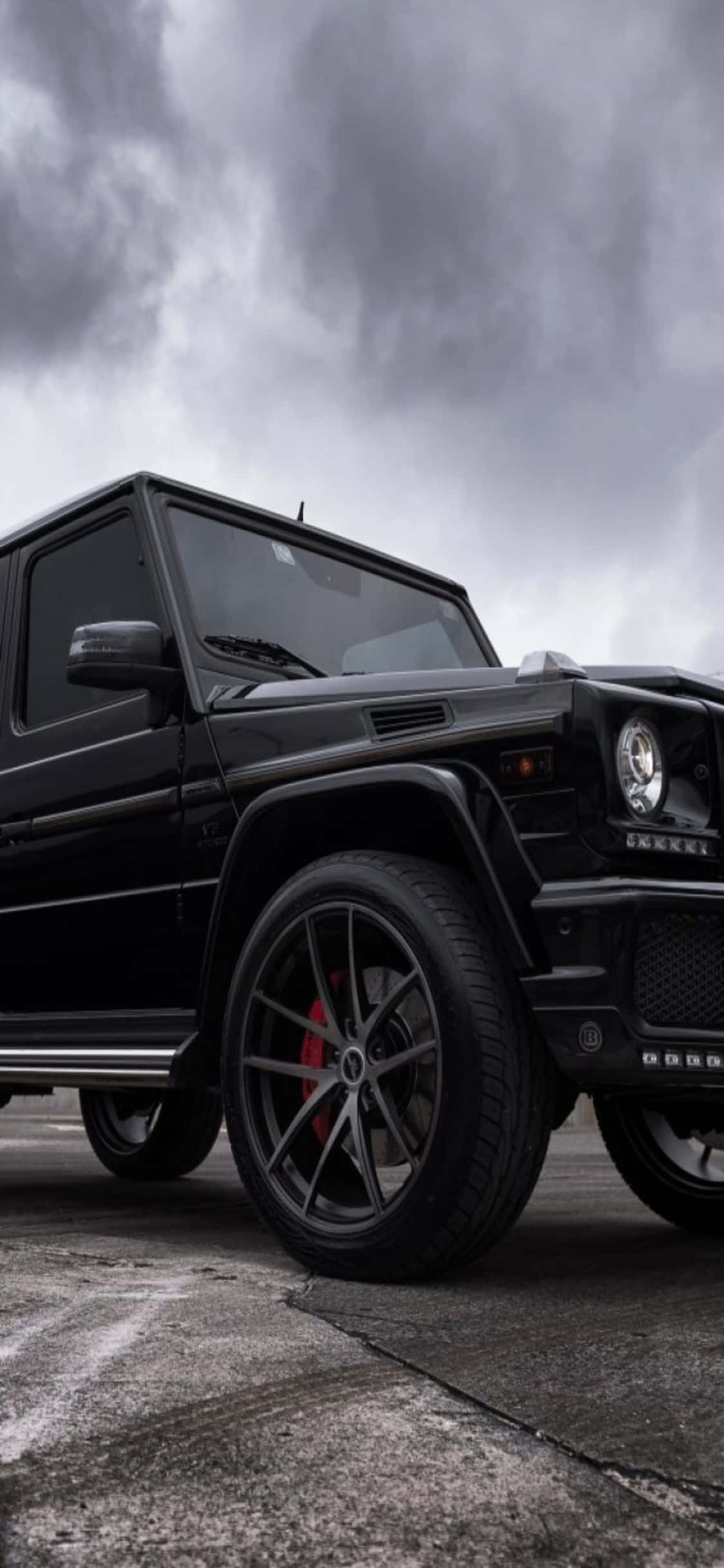 Mercedes 桌布 壁紙 — 一輛時尚的黑色Mercedes G-Wagen配備紅色制動卡鉗，停在風暴灰色天空下的裂縫路面上。