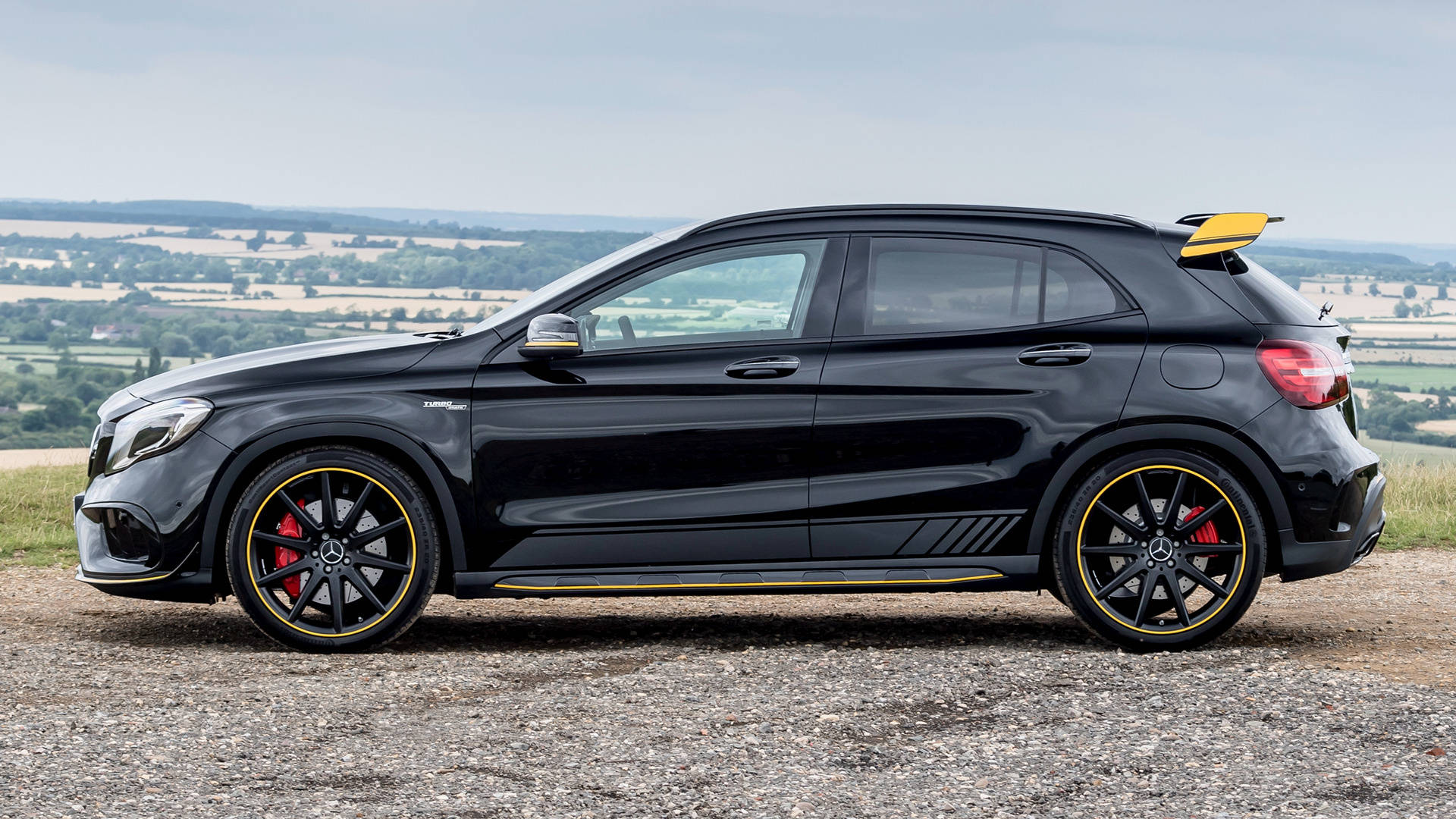 amg iPhone 桌布 壁紙 — 一輛時尚的黑色Mercedes-AMG GLA45 S停在碎石上，背景是起伏的鄉村景觀。