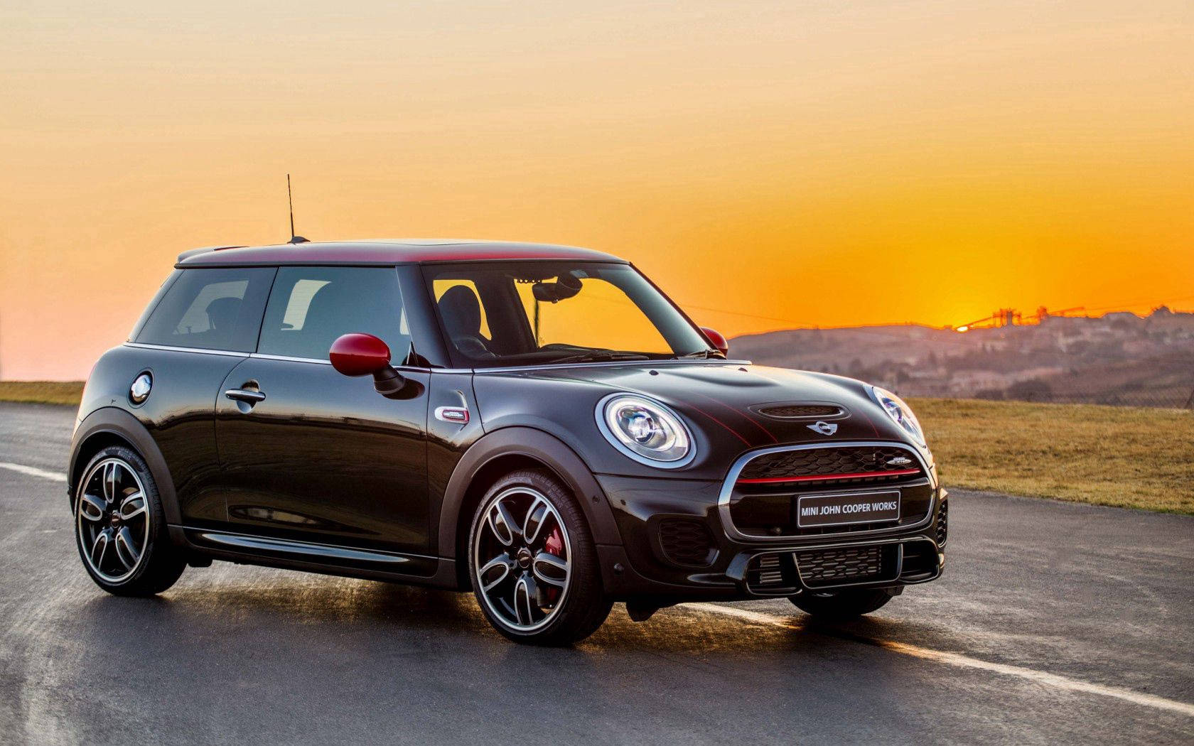 Mini Cooper 桌布 壁紙 — 一輛時尚的黑色MINI John Cooper Works跑車在金色日落和溫暖橙色天空下的開闊道路上閃閃發光。