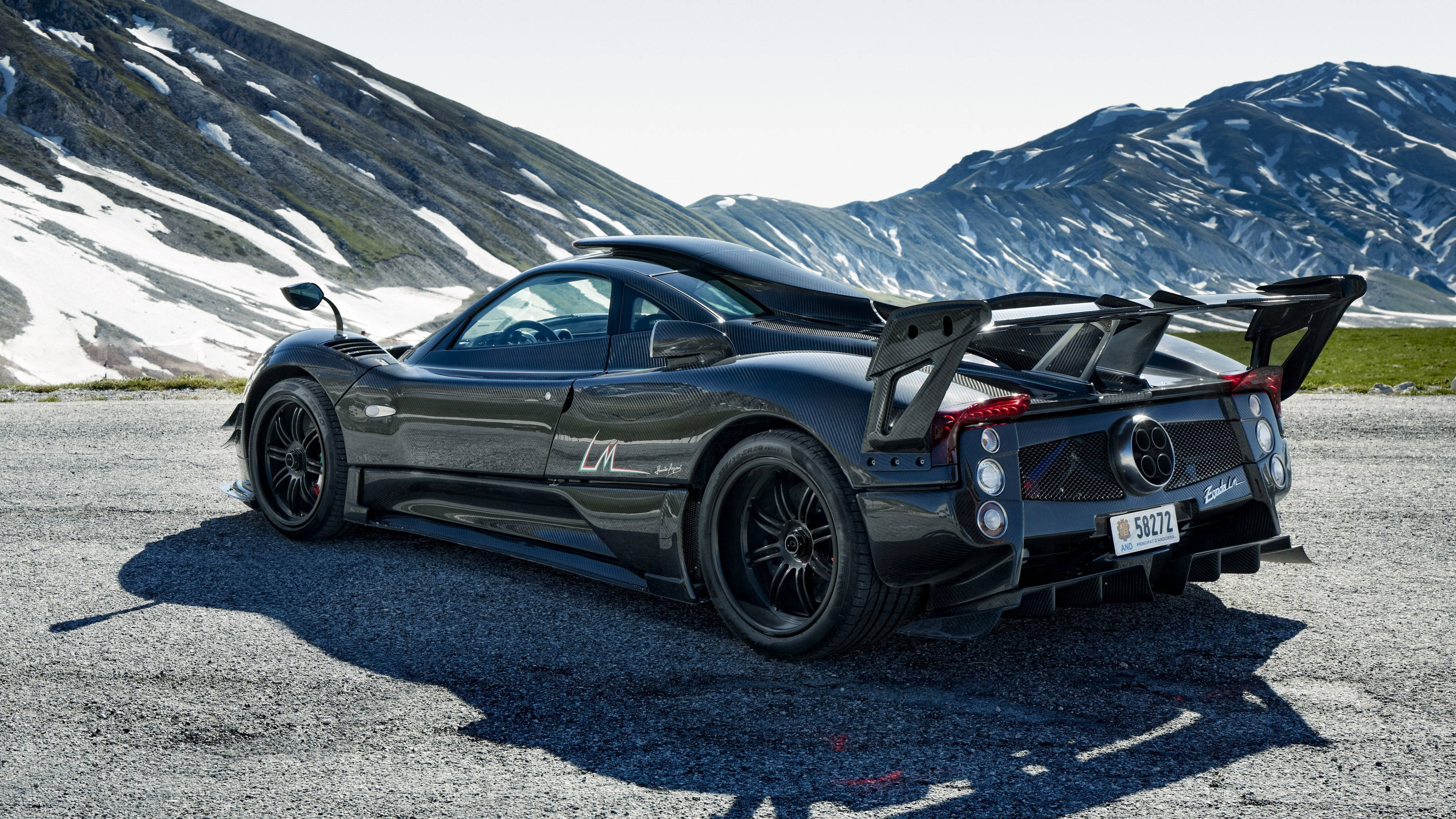 Pagani 桌布 壁紙 — 一輛時尚的黑色Pagani Huayra停在碎石路上，背後有白雪覆蓋的山脈在明亮的日光下聳立。
