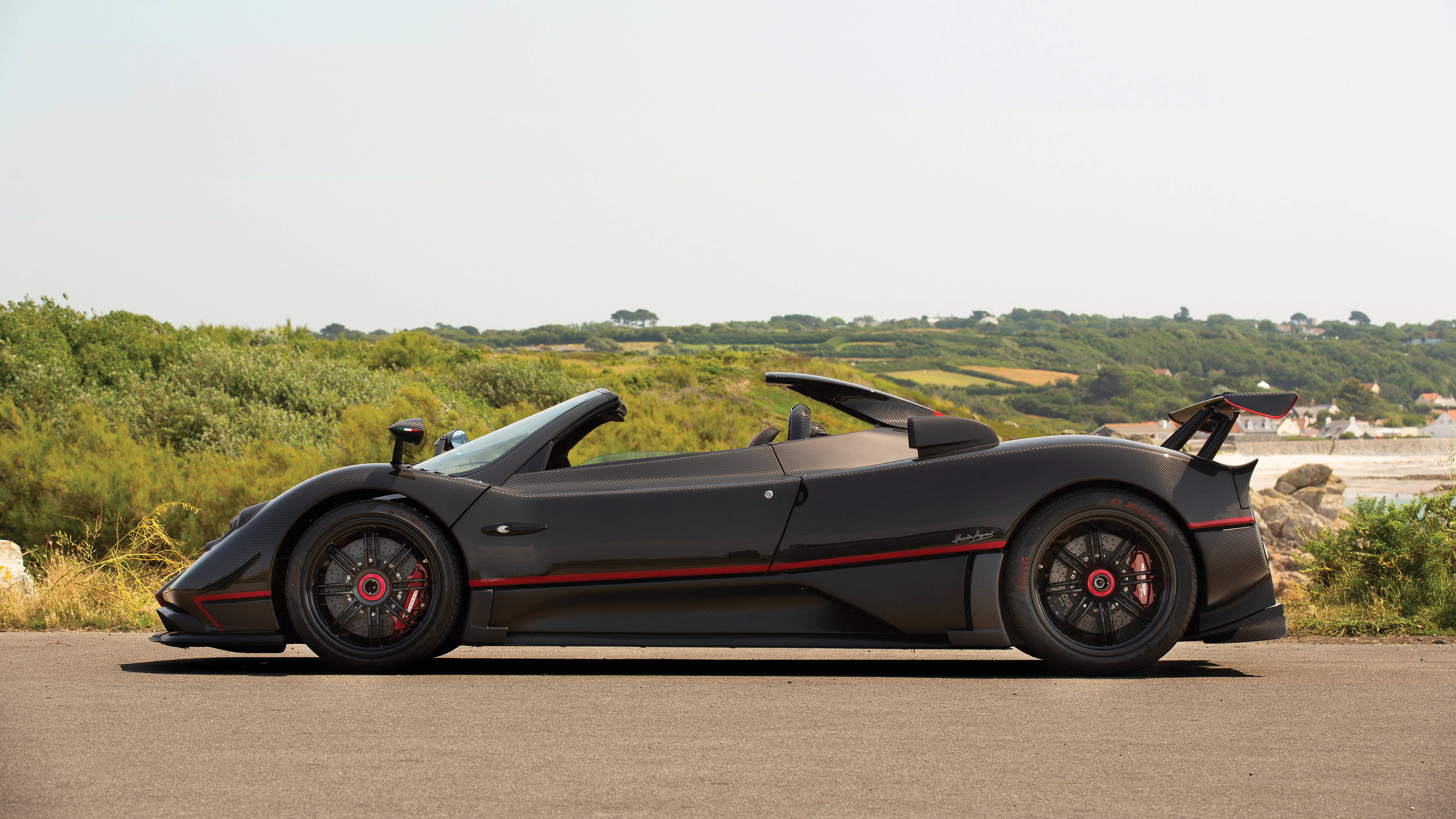 pagani iPhone 桌布 壁紙 — 一輛時尚的黑色Pagani Zonda Cinque Roadster，帶有紅色點綴，停泊在明亮天空下綠丘旁的沿海道路上。