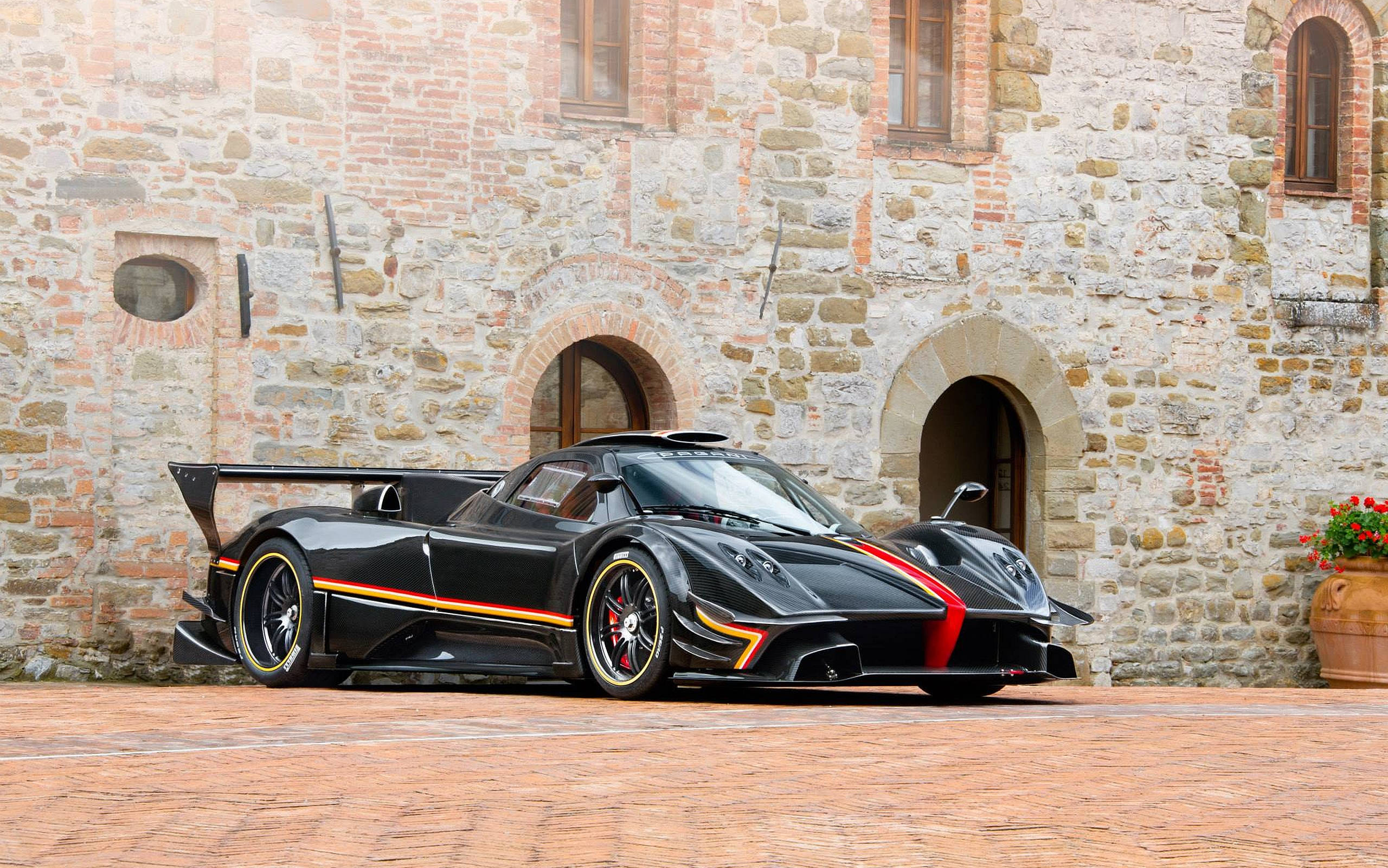pagani iPhone 桌布 壁紙 — 一輛時尚的黑色Pagani超跑，帶有紅色和金色點綴，停在磚砌庭院前，背景是質樸的石頭建築。