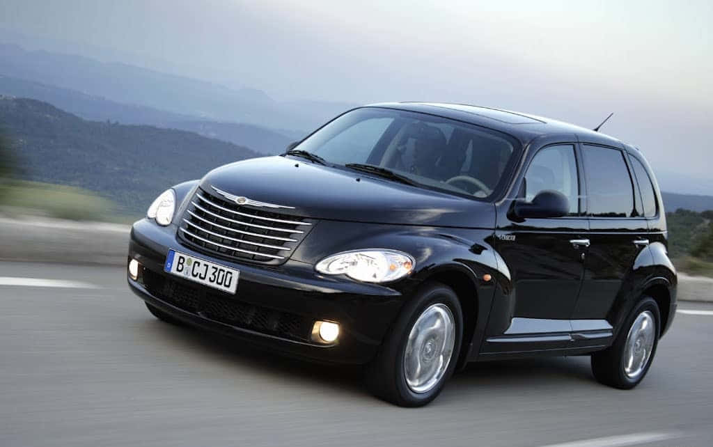 Chrysler Pt Cruiser 桌布 壁紙 — 一輛時尚的黑色PT Cruiser沿著蜿蜒的山路行駛，背景是模糊的綠色山丘。