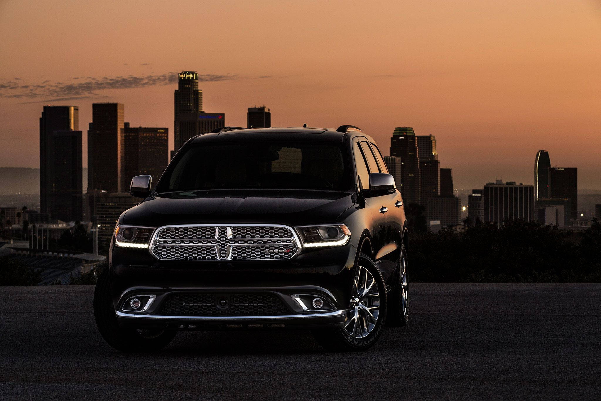 Dodge 桌布 壁紙 — 一輛時尚的黑色SUV停泊在山上，俯瞰夕陽下溫暖橙色和粉紅色調的城市天際線。
