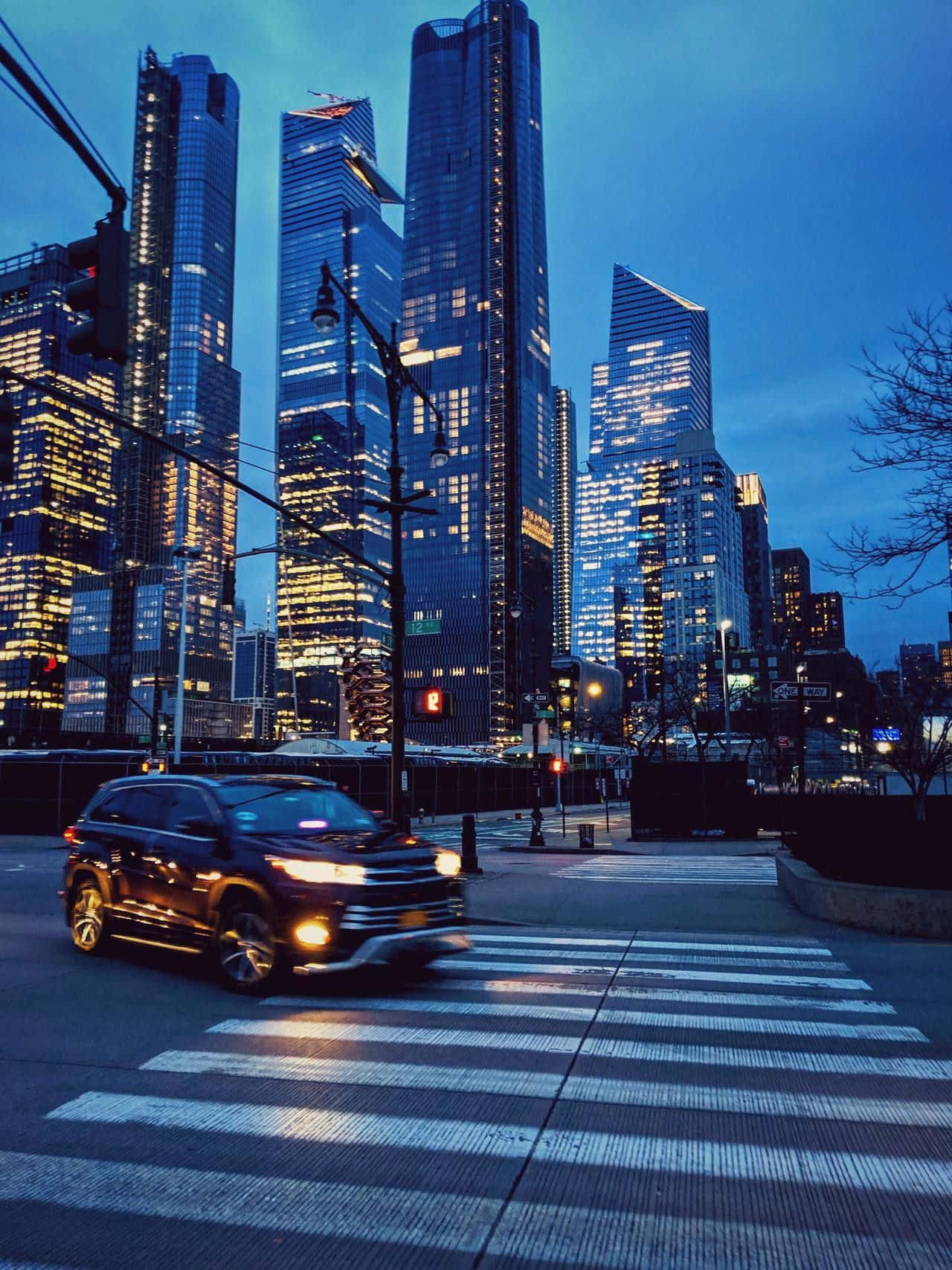 Nyc Aesthetic 桌布 壁紙 — 一輛時尚的黑色SUV在暮色天空下，穿過發光的行人穿越道，背後是高聳的玻璃摩天大樓。