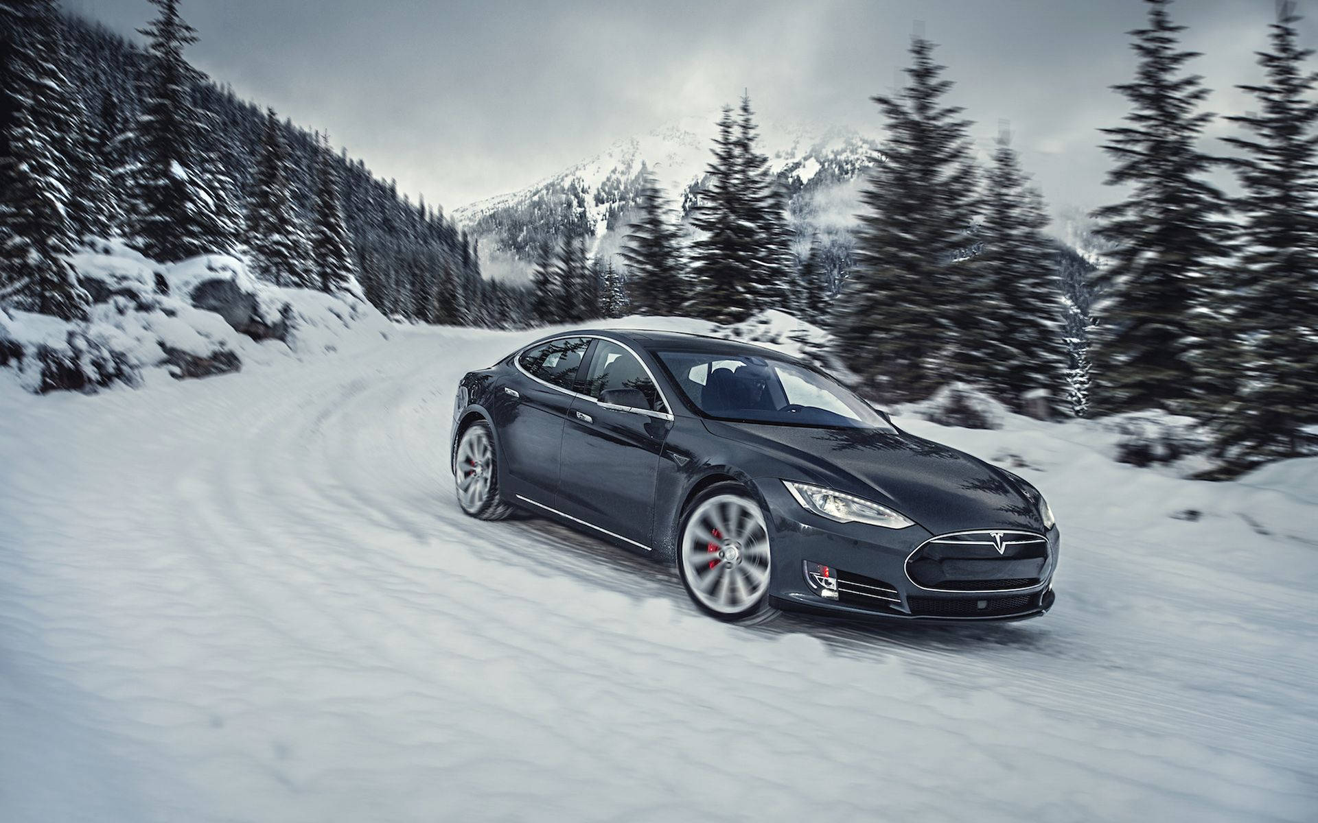 特斯拉 桌布 壁紙 — 一輛時尚的黑色Tesla Model S在陰天下行駛於蜿蜒的積雪山路，兩旁是常青樹。