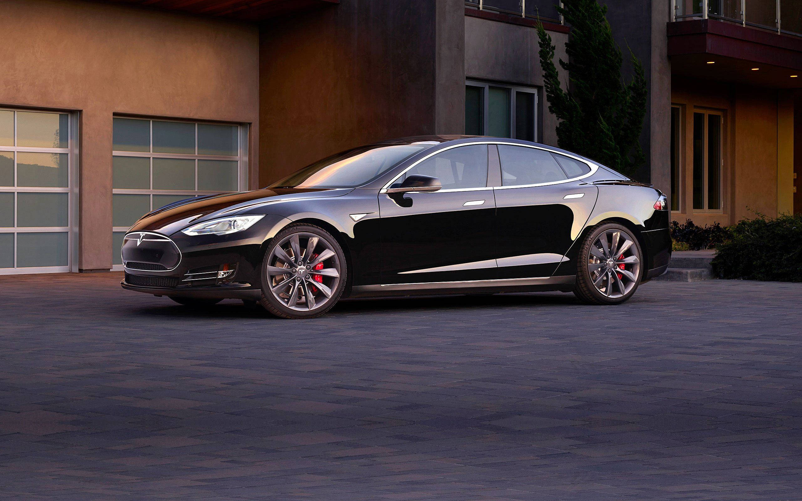 特斯拉 桌布 壁紙 — 一輛時尚的黑色Tesla Model S停在鋪砌的車道上，前面是帶有溫暖照明的現代房屋。