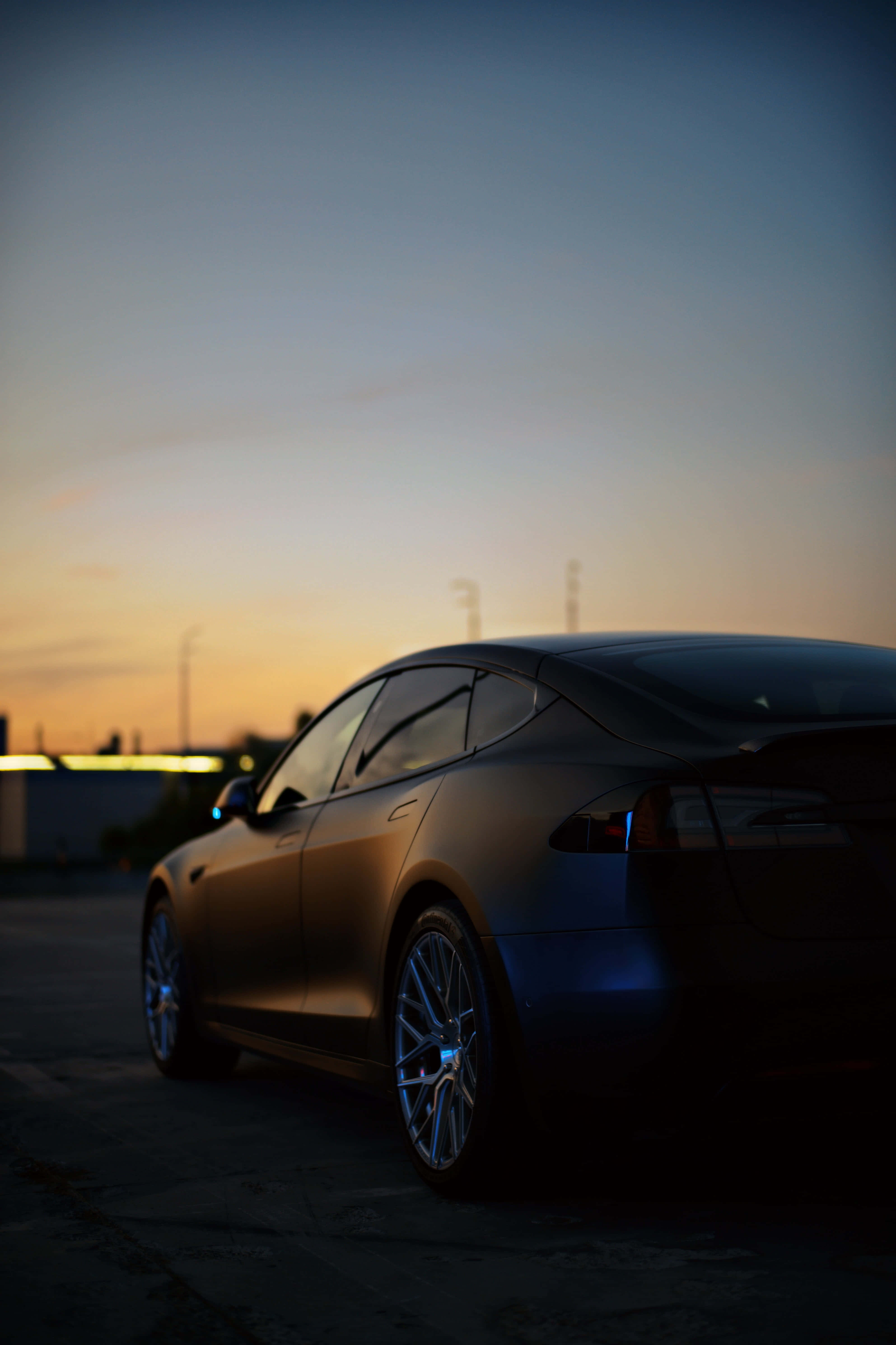 Tesla iPhone 桌布 壁紙 — 一輛時尚的黑色Tesla轎車在黃昏時停泊，溫暖的橙色天空反射在其光滑表面上。