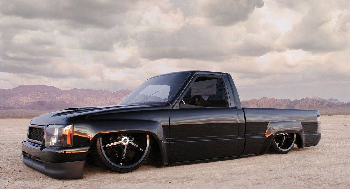 Trucks 桌布 壁紙 — 一輛時尚的黑色低趴皮卡車停在廣闊的沙漠中，背景是戲劇性的多雲天空和遠處的山脈。