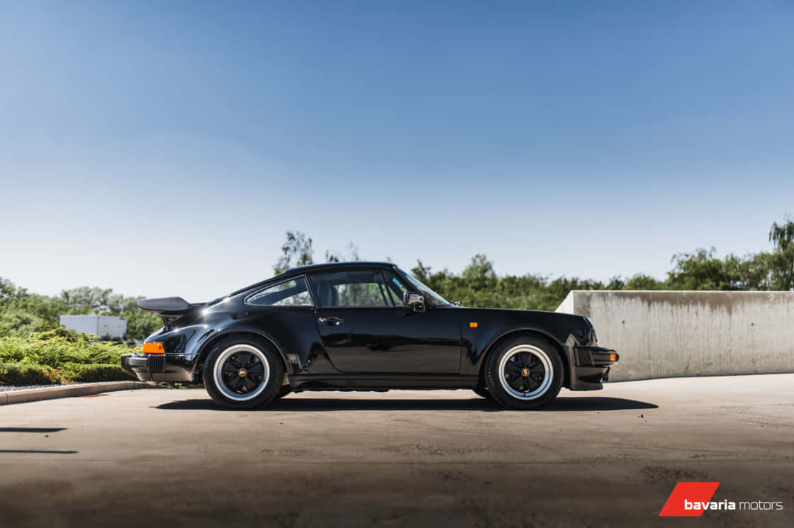 Porsche 930 桌布 壁紙 — 一輛時尚的黑色保時捷911 Turbo 停在陽光普照的混凝土表面上，背景有綠植和矮牆。