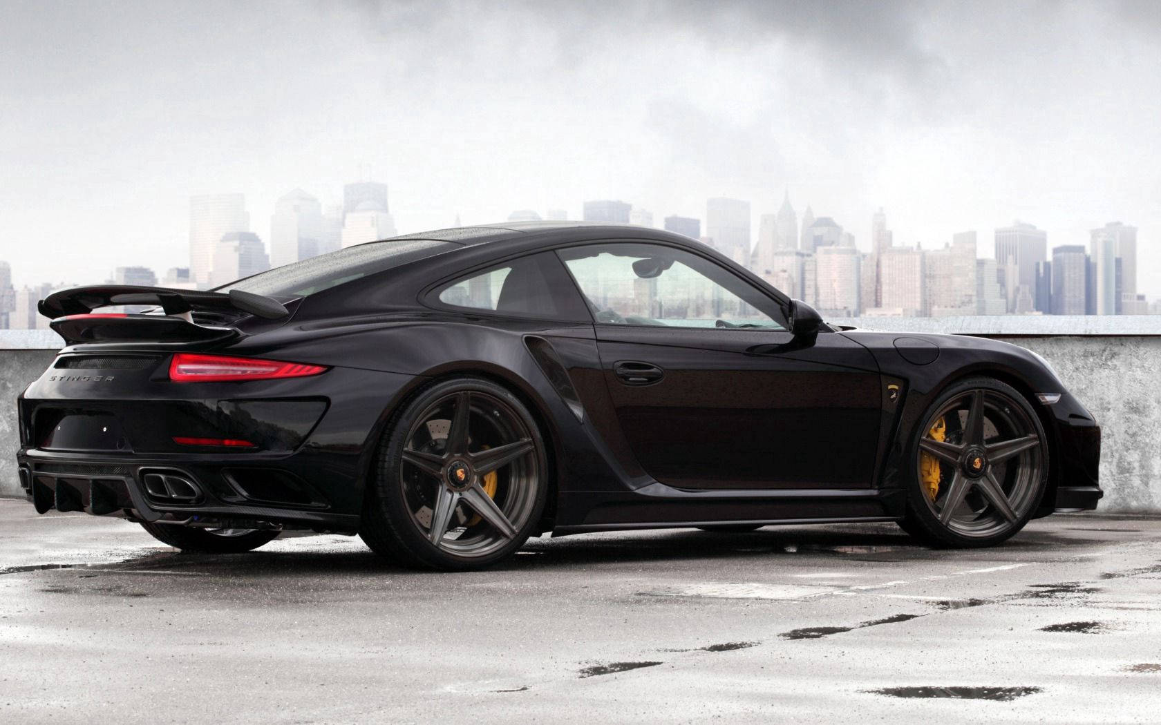保時捷 桌布 壁紙 — 一輛時尚的黑色保時捷911 Turbo S，配備金色制動卡鉗，停在濕漉漉的屋頂上，背景是朦朧的城市天際線。